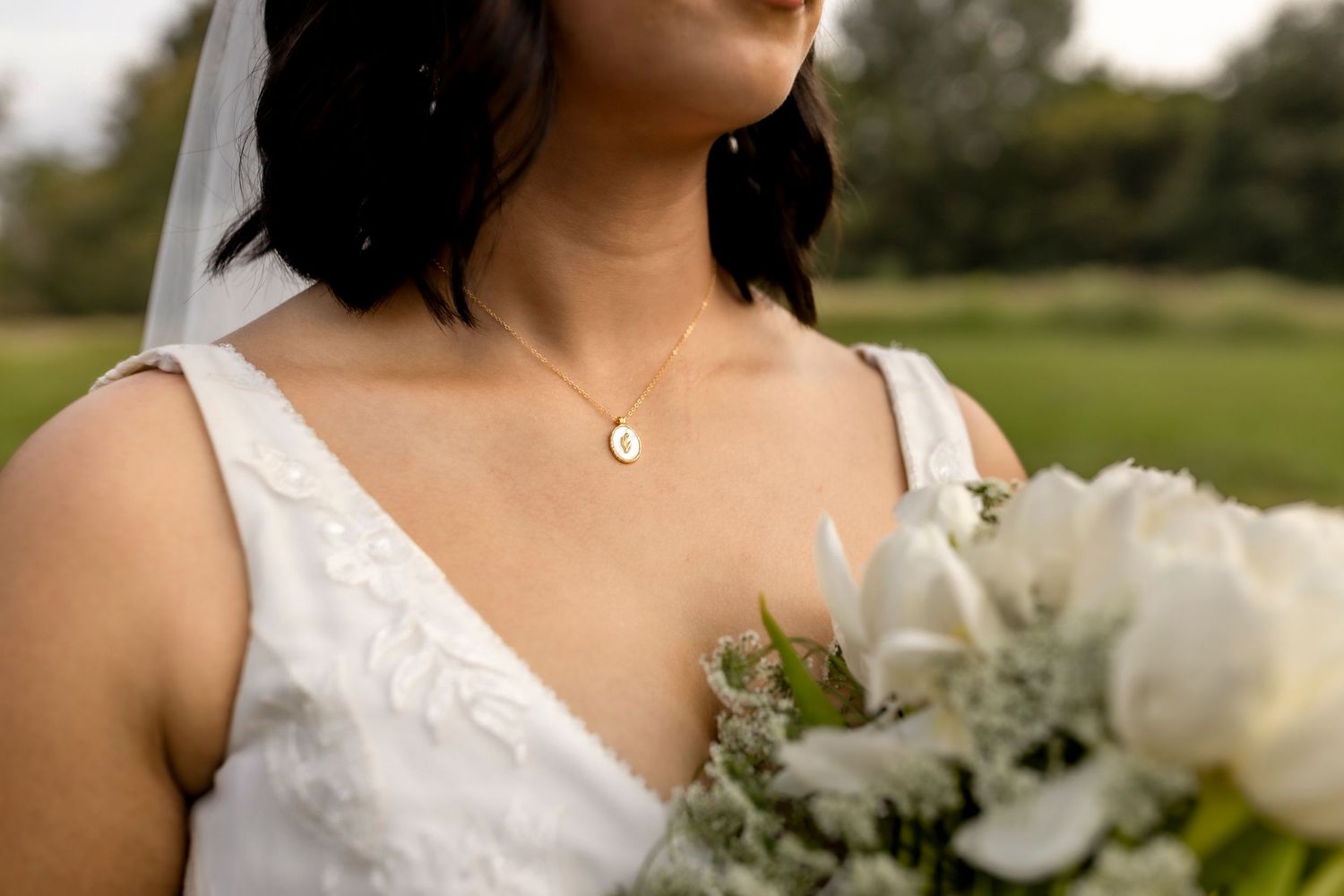 Close-up of bride in white V-neck gown wearing a delicate gold oval necklace, holding white tulip bouquet.