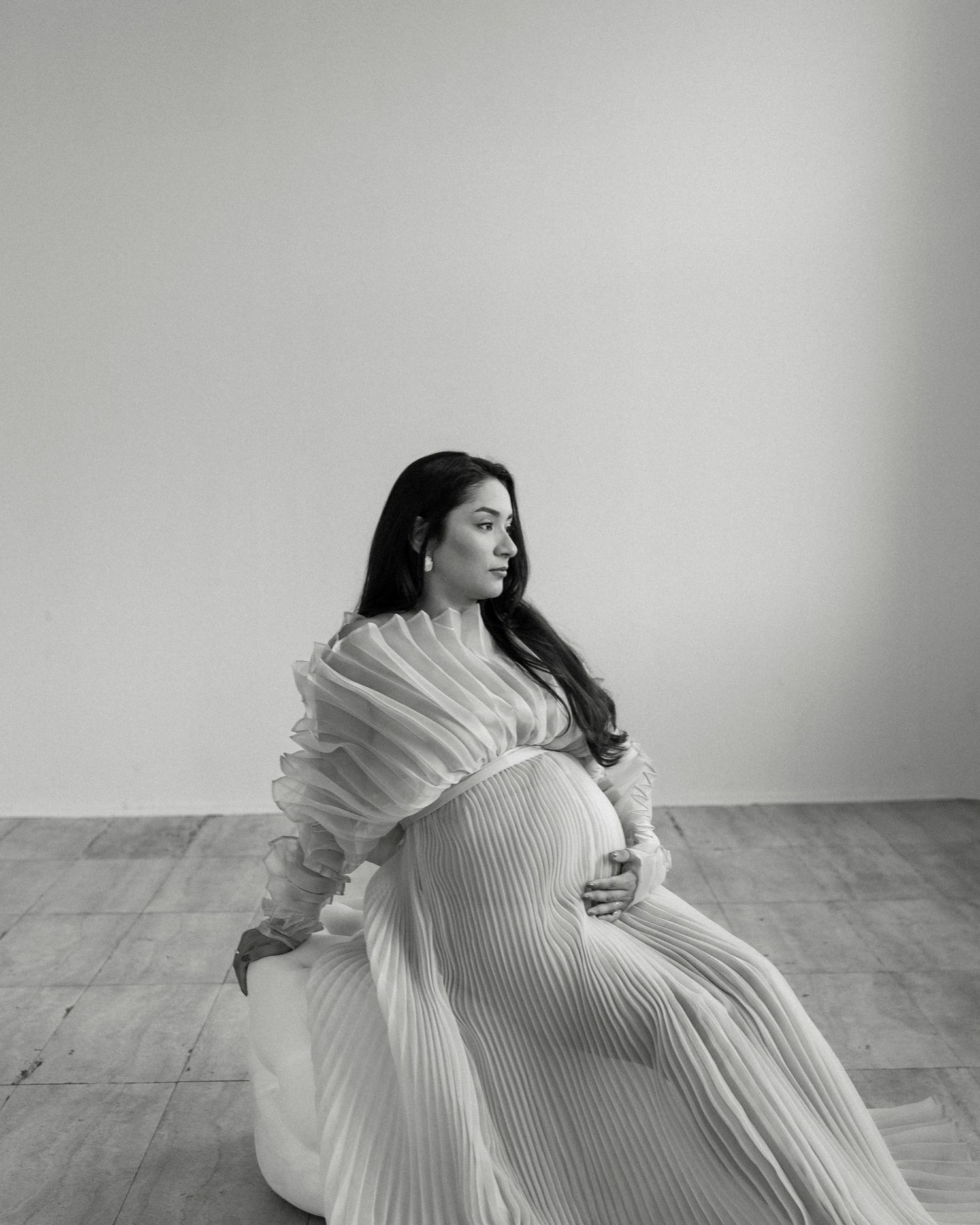 Black and white maternity photo of pregnant woman in pleated white gown sitting gracefully on studio floor.