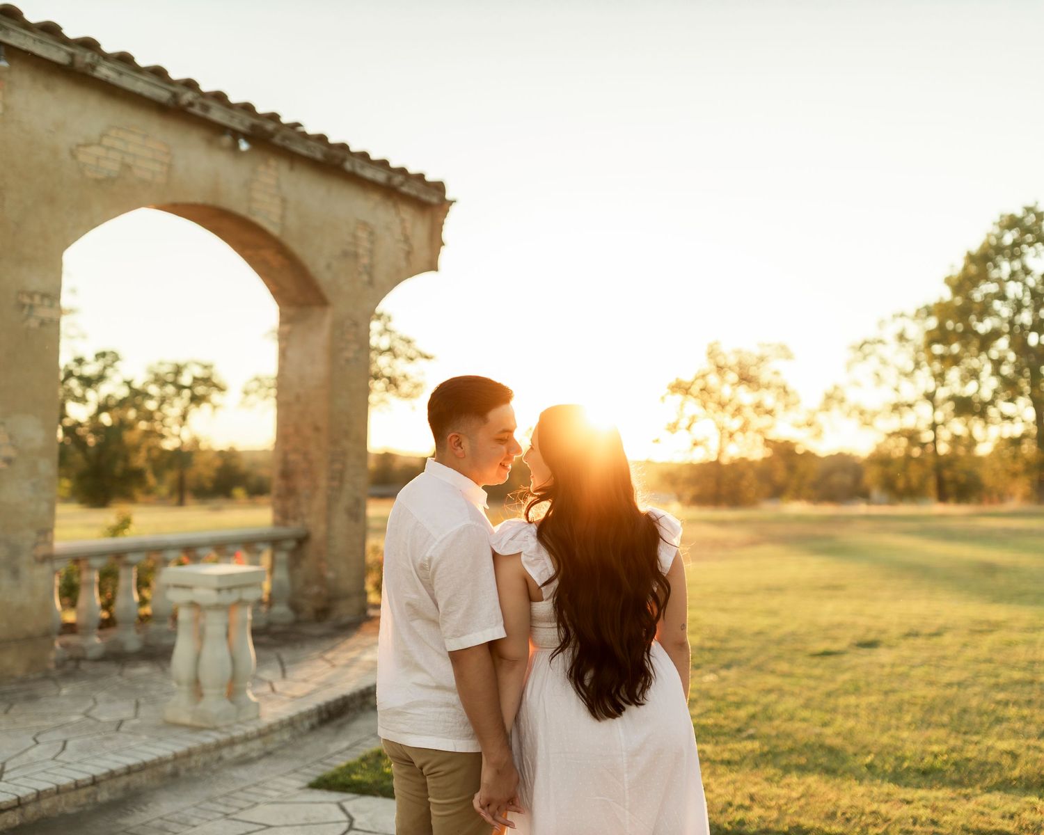 Couple sharing a golden sunset kiss near a stone arch in a lush green garden.