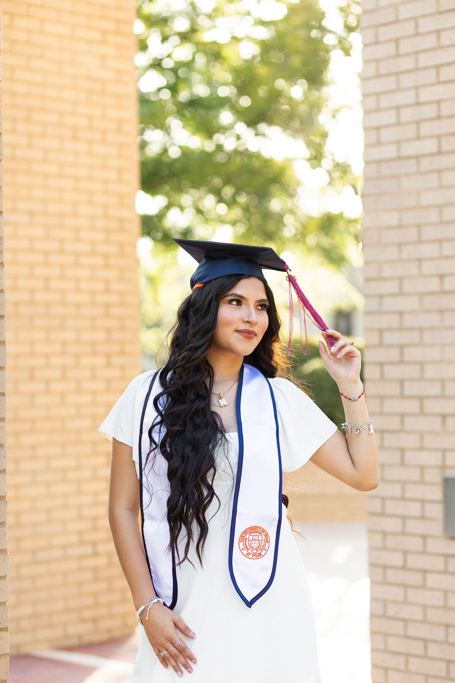 Female college graduate in white gown and black cap holding tassel outdoors on campus.