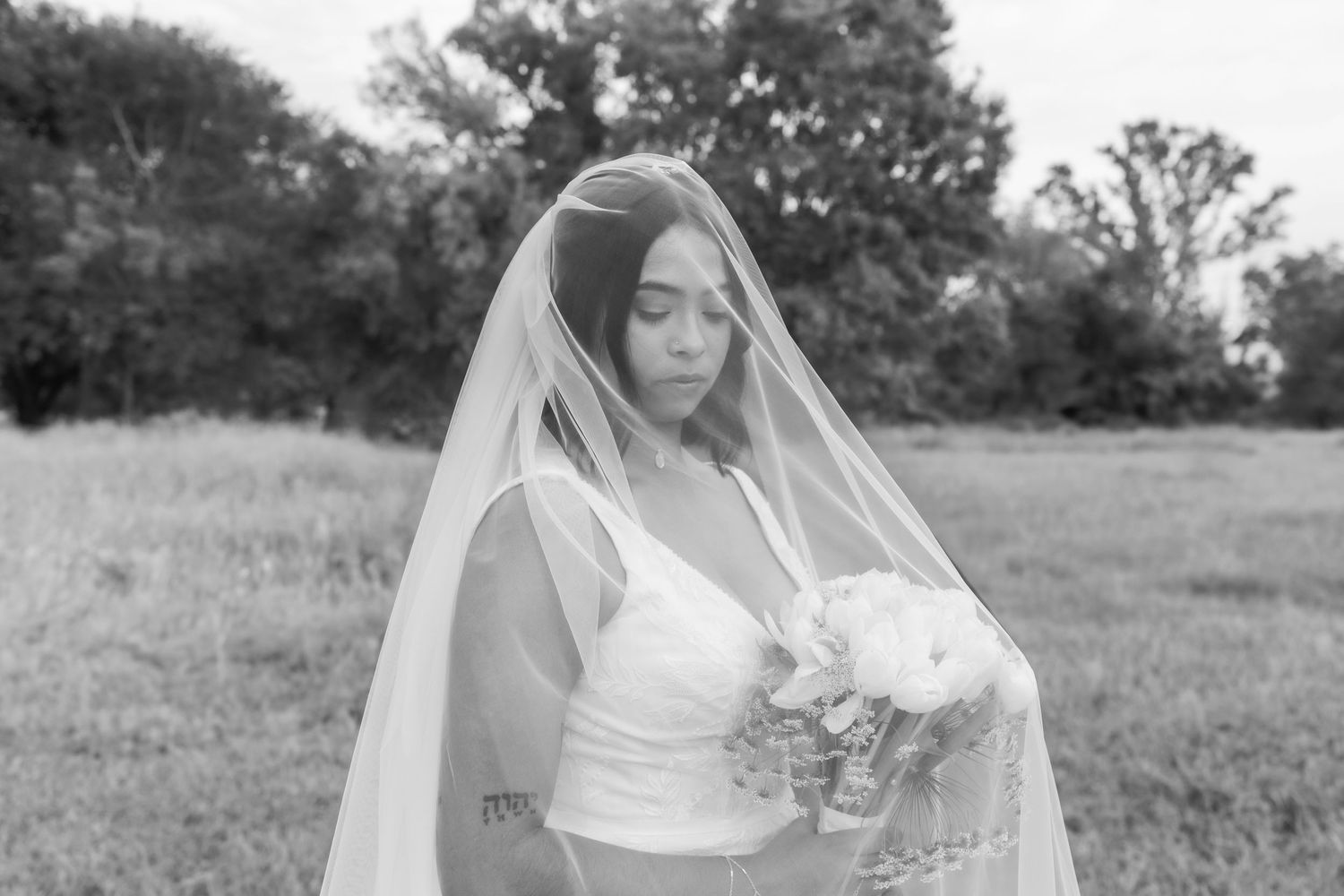 Black and white bridal portrait of bride covered by veil holding bouquet in outdoor field setting.