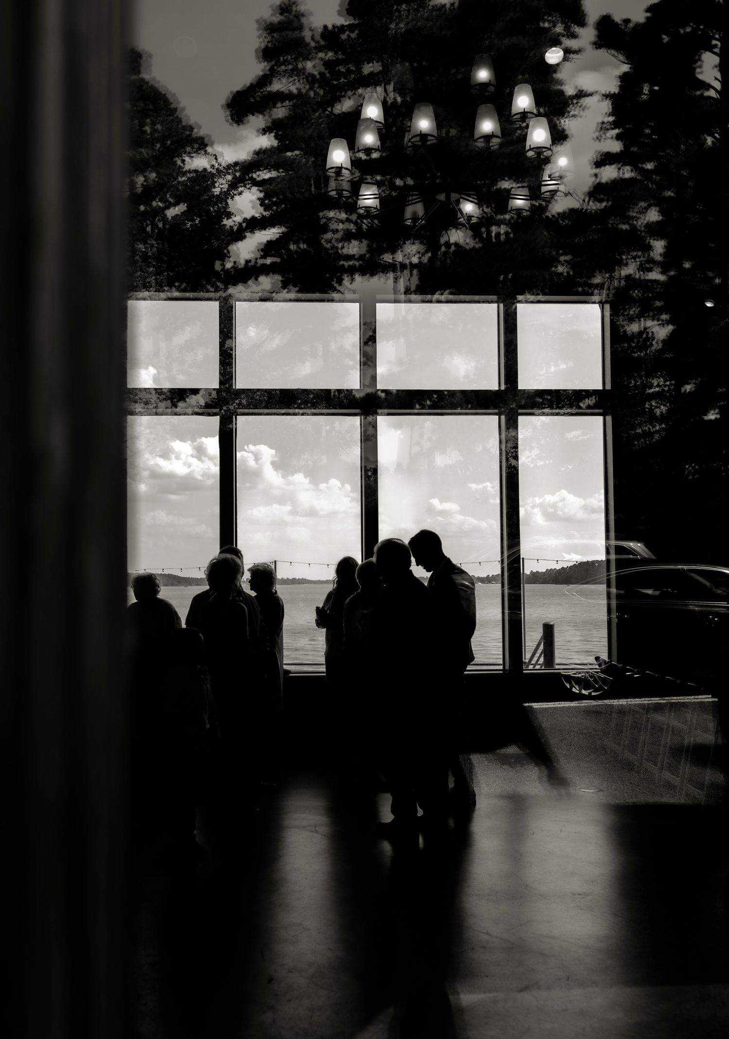 Silhouettes of wedding guests gathered near large windows with a scenic lake view in black and white.