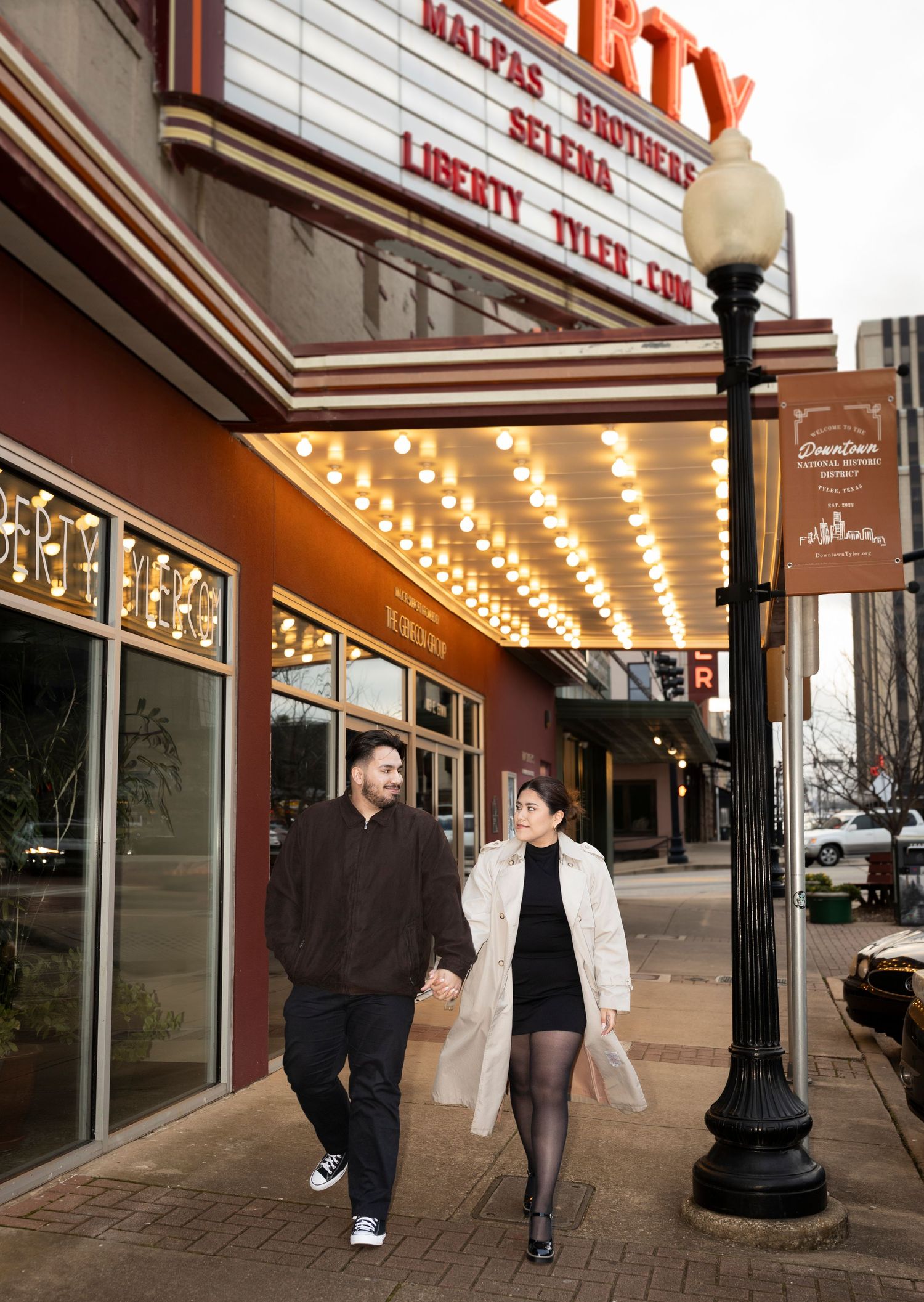 A couple walks hand in hand past the Liberty Theater marquee on a downtown city sidewalk at night.