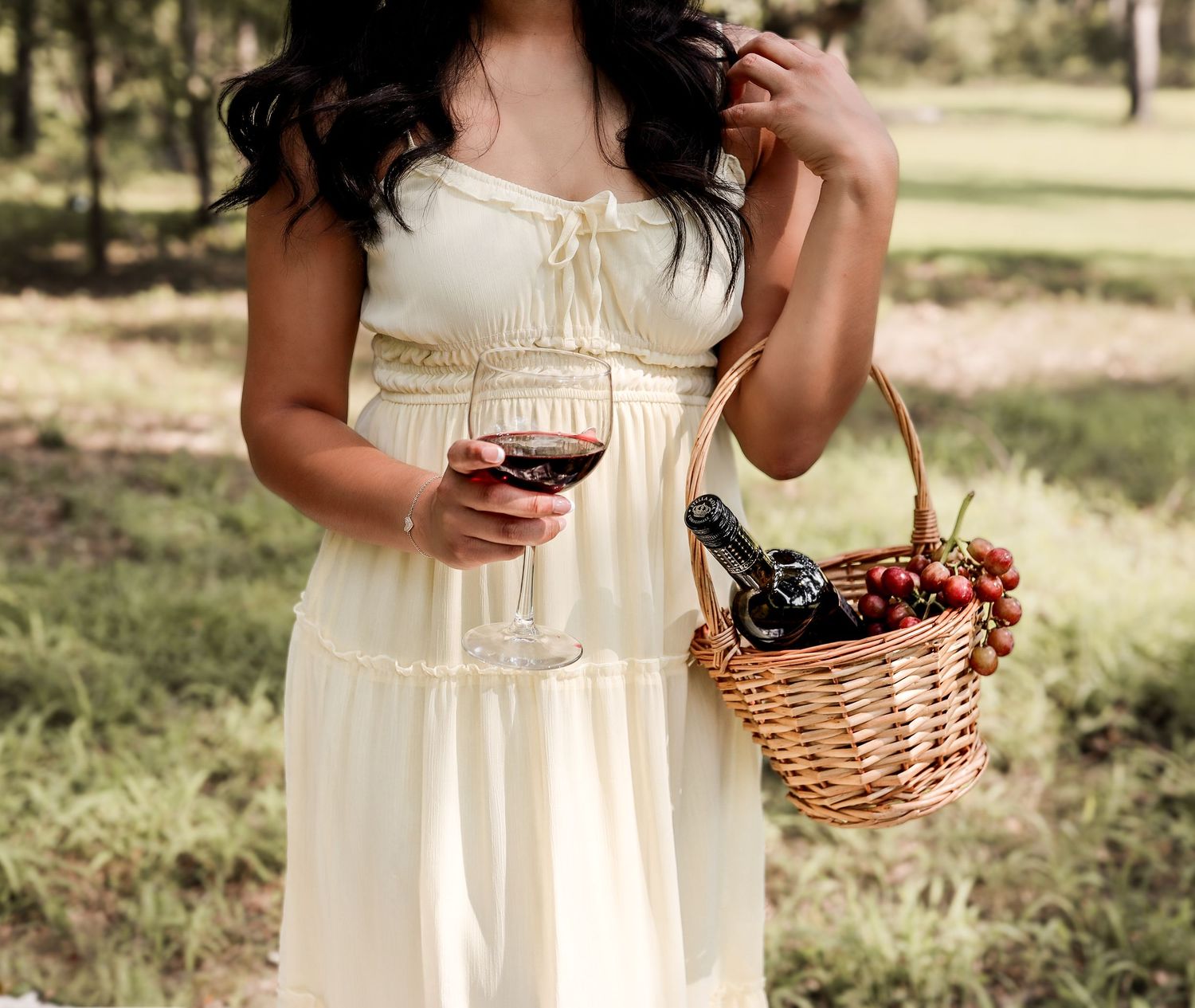 Woman in cream dress holding wine glass and wicker basket with cherries and wine bottle at outdoor picnic.