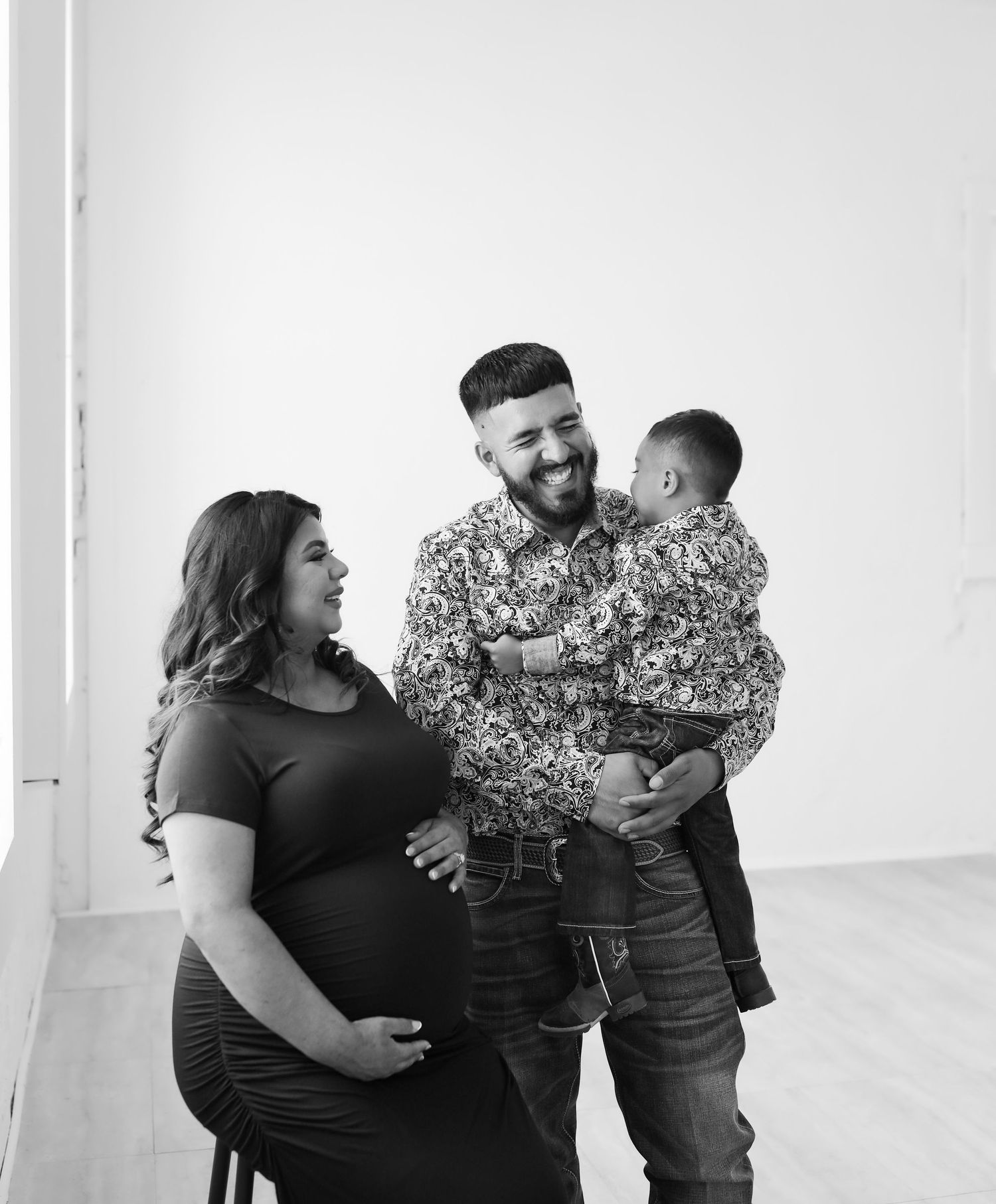 Black and white photo of a pregnant woman smiling at her partner holding their toddler son.
