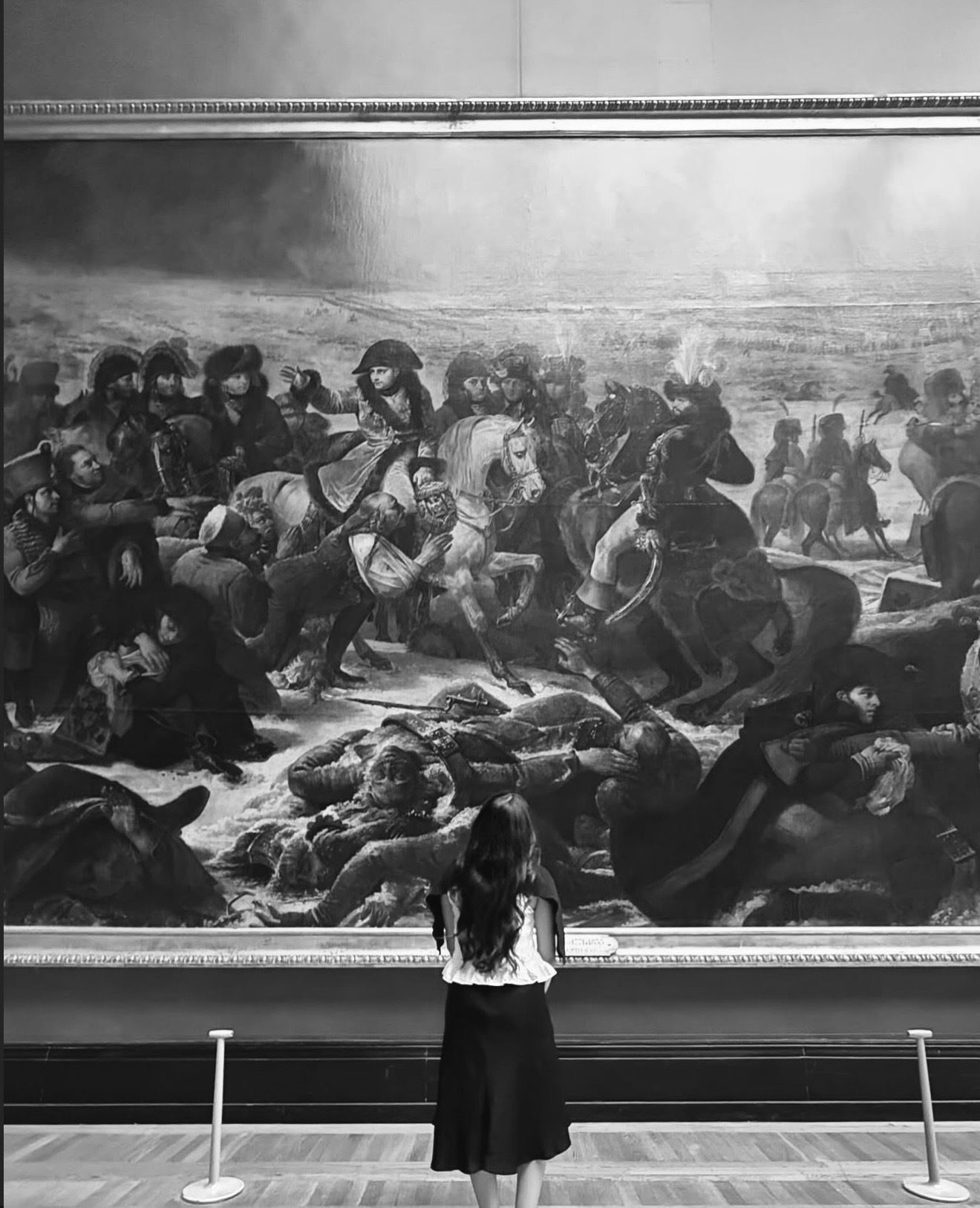 Black and white photo of a woman viewing a large battle painting in a museum gallery.