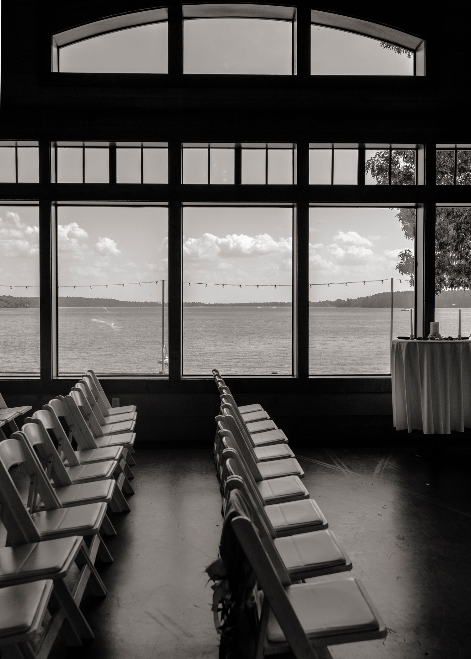 Empty wedding ceremony chairs arranged in rows before large windows with scenic lake view.