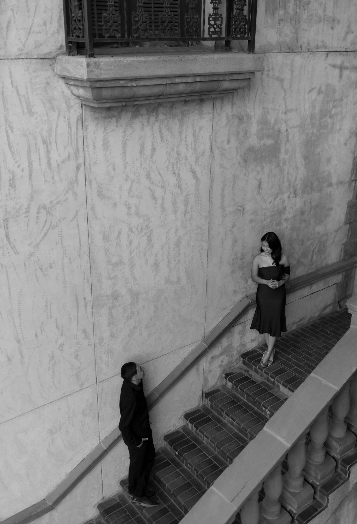 Black and white photo of couple on grand staircase beside tall marble wall.