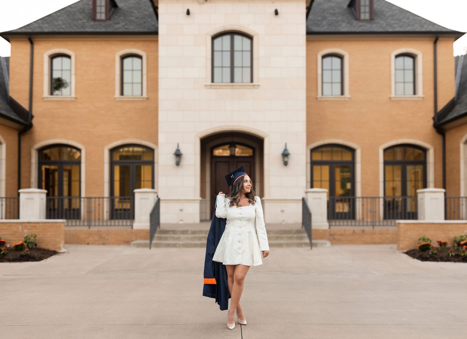 Graduate in white dress carries navy regalia while walking in front of elegant tan stone mansion.