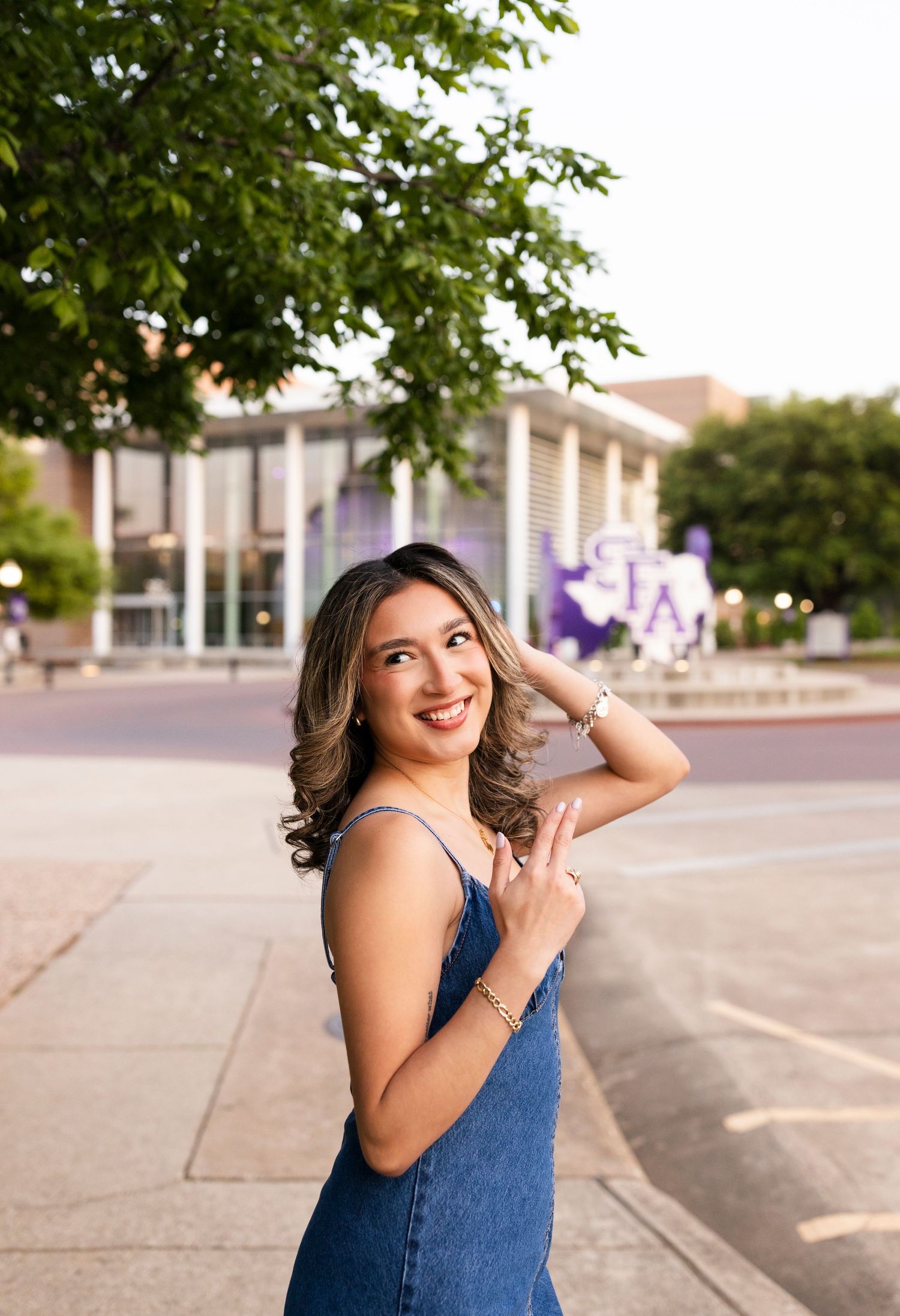 Smiling young woman in denim dress poses outdoors near SFA university sign during golden hour senior photos.