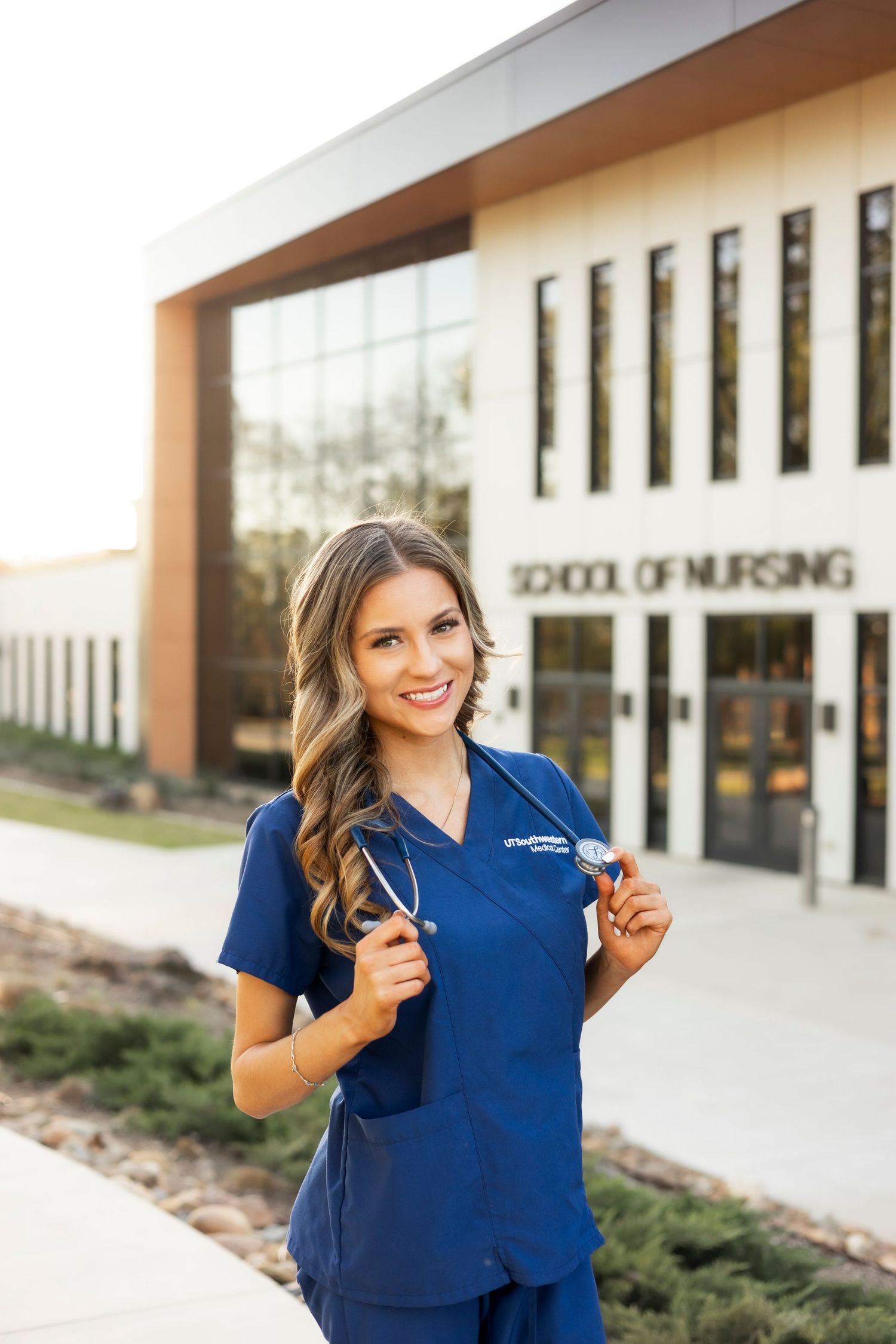 Smiling nursing student in blue scrubs holds stethoscope outside School of Nursing building.