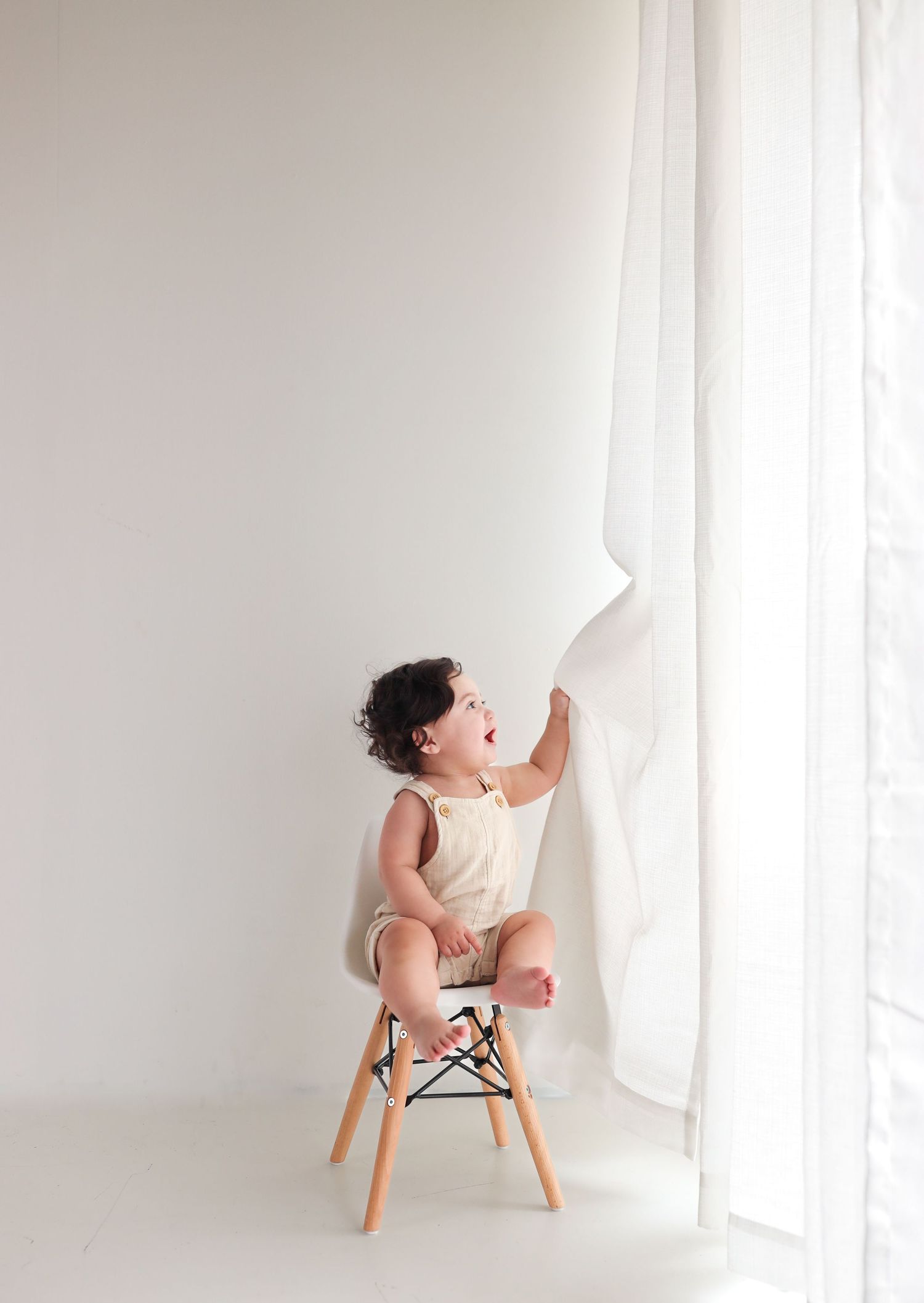 Joyful curly-haired toddler in white romper sitting on small chair, pulling sheer curtain in bright white studio.