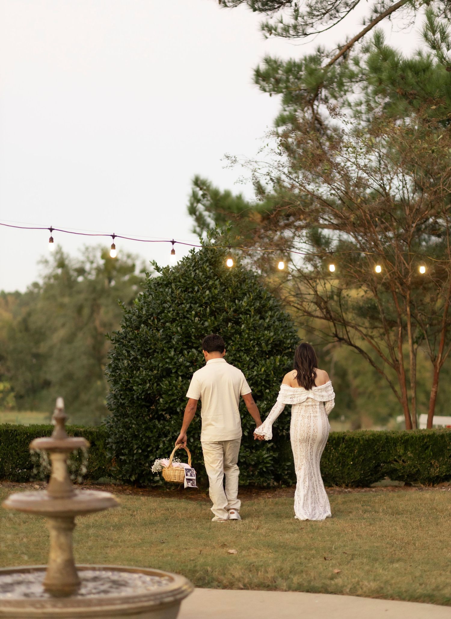 Couple walking hand in hand through a garden with string lights and a stone fountain.