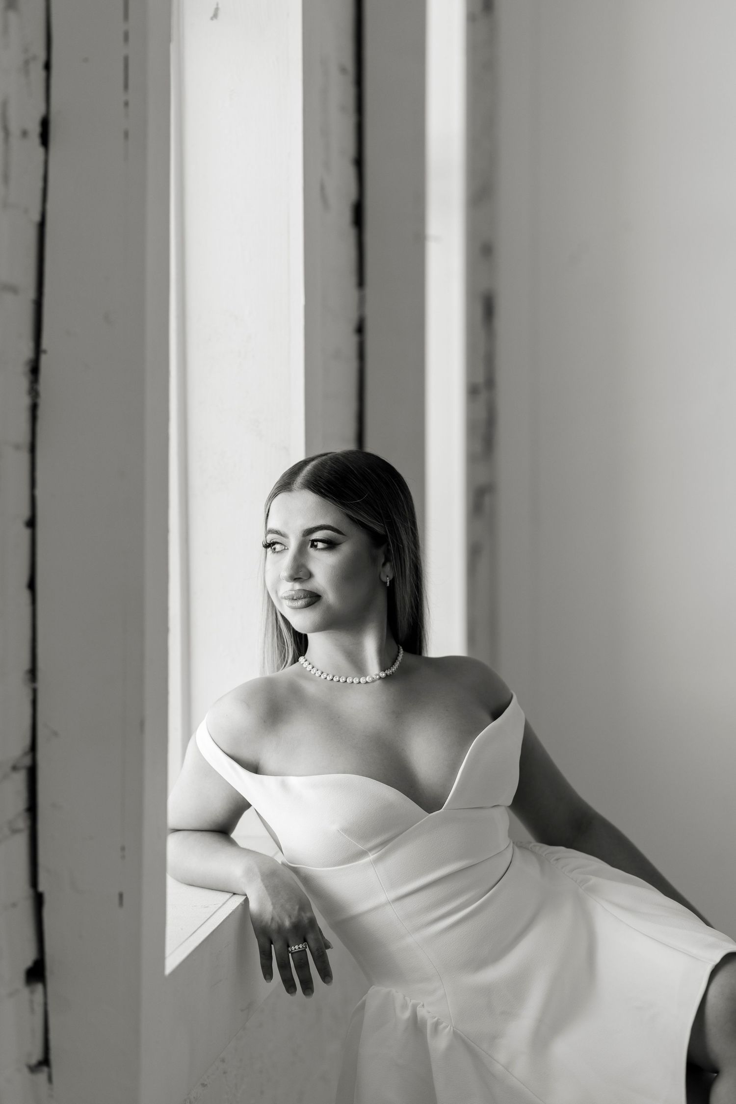 Black and white portrait of elegant woman in off-shoulder white gown leaning on windowsill.