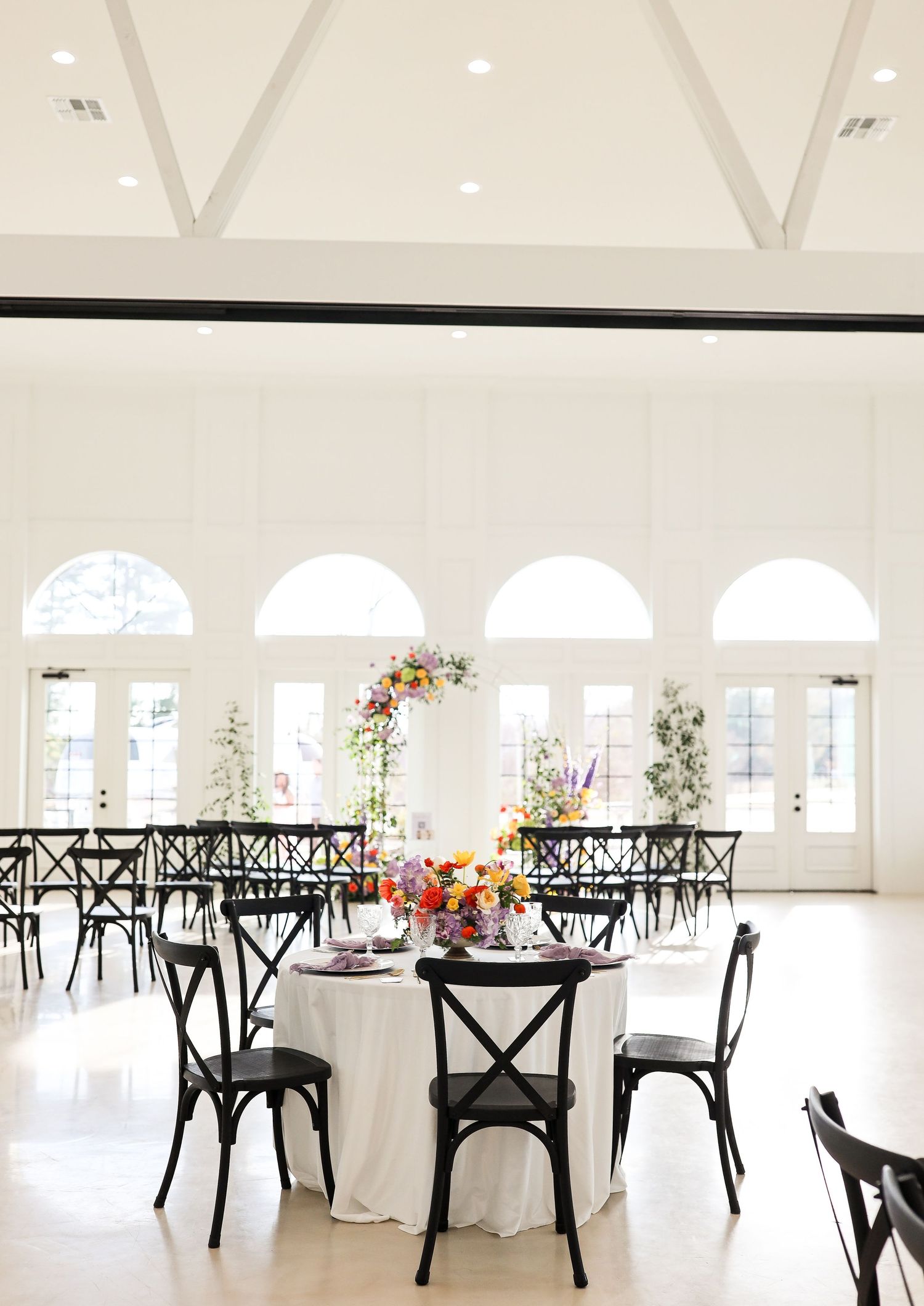 Bright white wedding reception venue with black crossback chairs, arched windows, and vibrant floral arrangements.