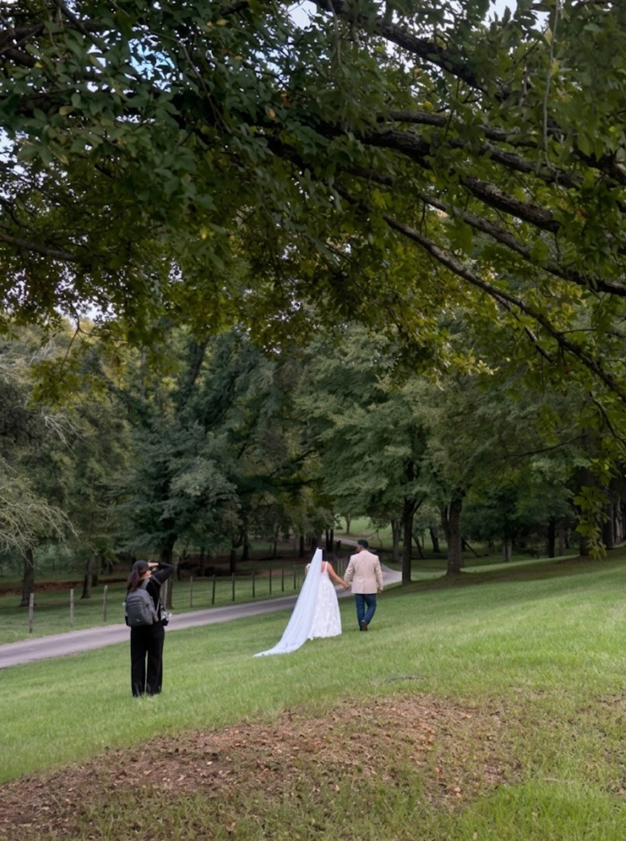 Photographer documenting bride and groom holding hands while walking through a scenic green tree-lined park.