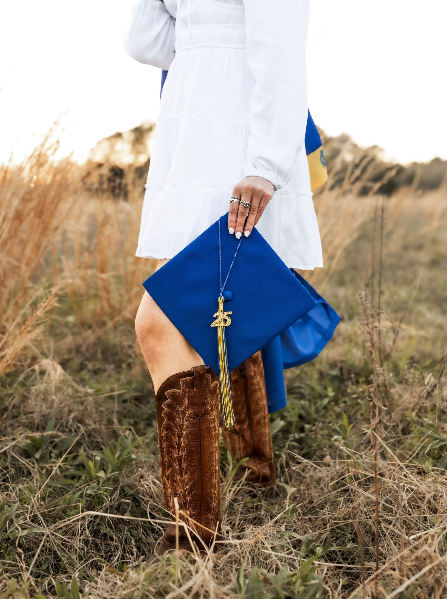 Graduate holding royal blue cap with gold 2025 tassel wearing white dress and brown cowboy boots in field.