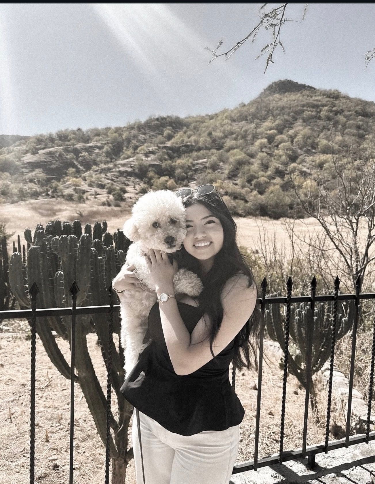 Smiling woman holding fluffy white poodle outdoors with desert hillside landscape in black and white.