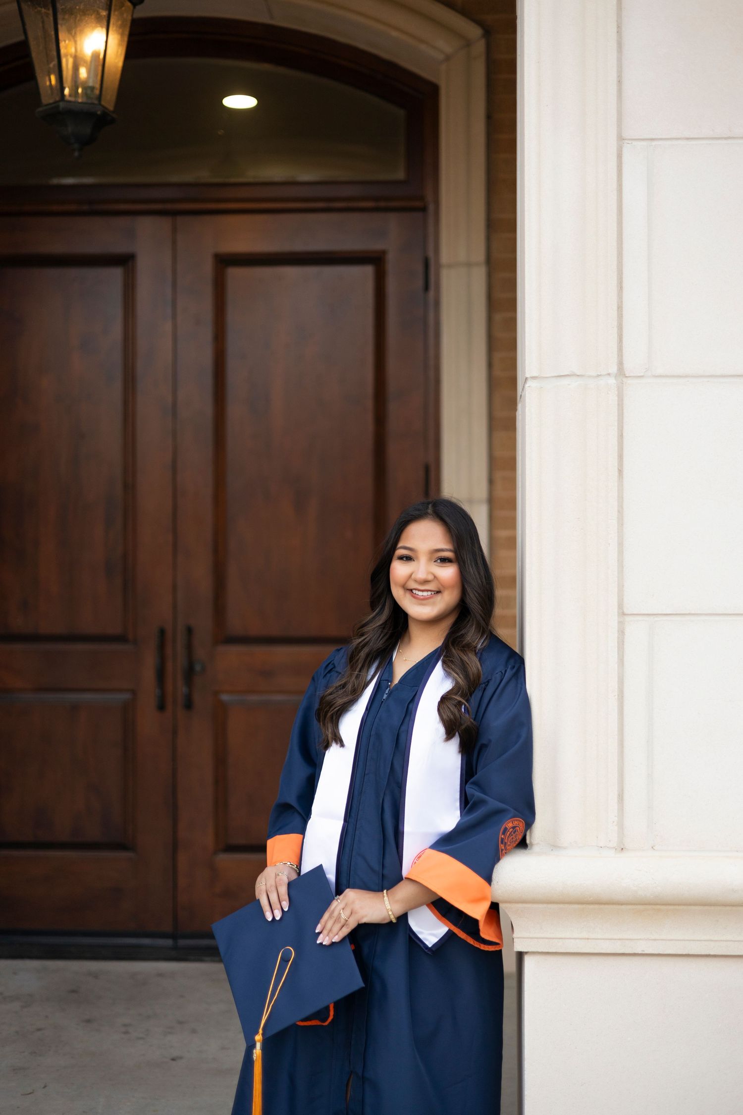 Female graduate in navy and orange cap and gown poses proudly in front of elegant wooden doors.