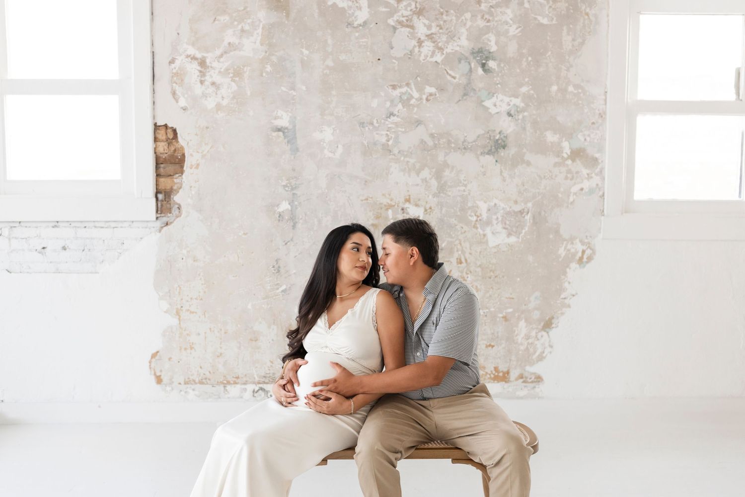 Couple sitting together on a bench, man kissing pregnant womans cheek in bright studio.
