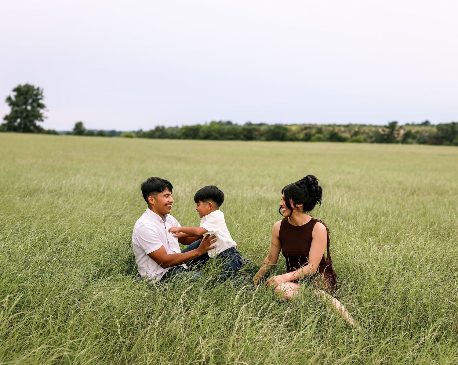 Family of three sitting together in tall green grass field, parents engaging playfully with their toddler on overcast day.