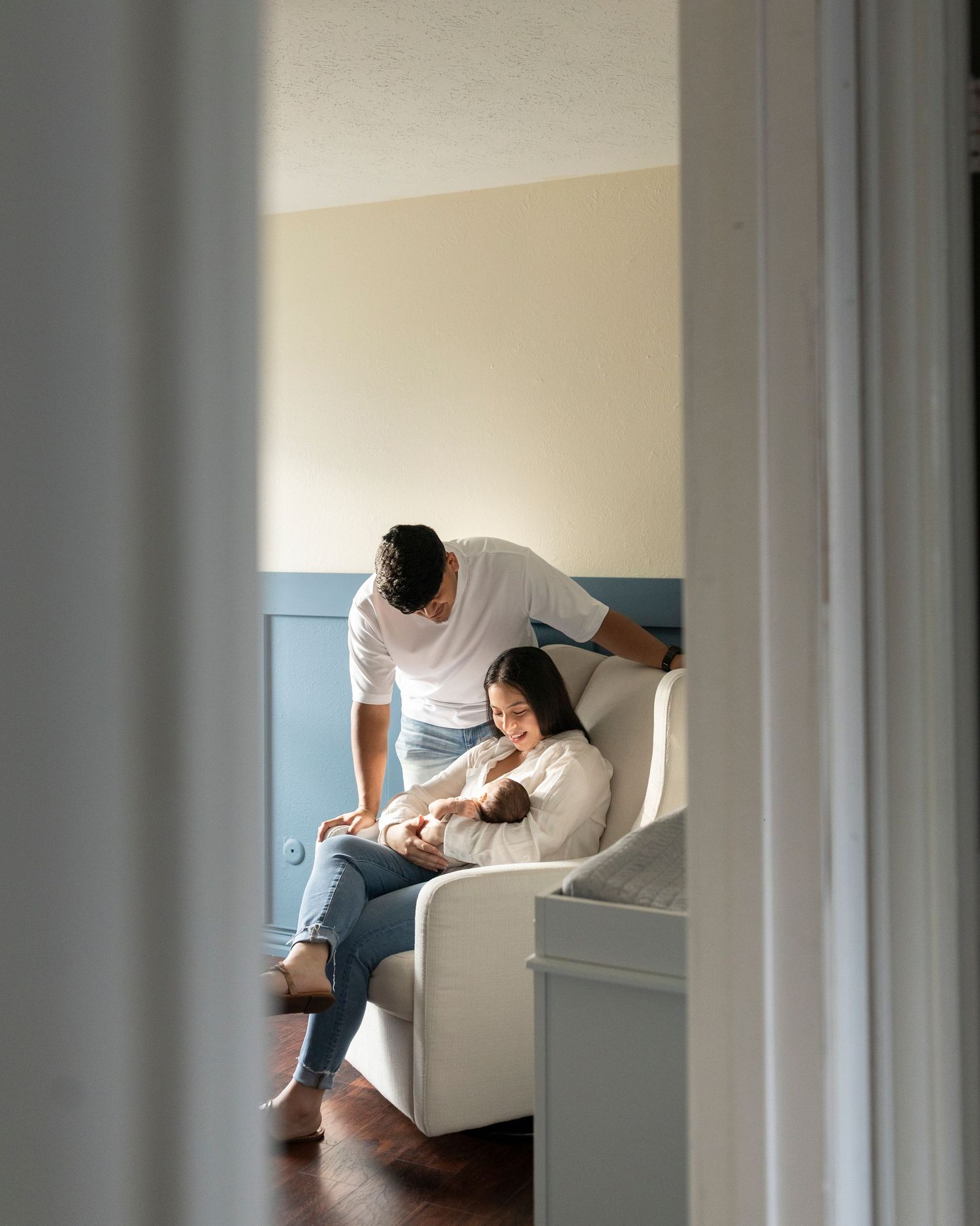 New parents in a softly lit nursery, mother seated holding newborn while father leans in close.