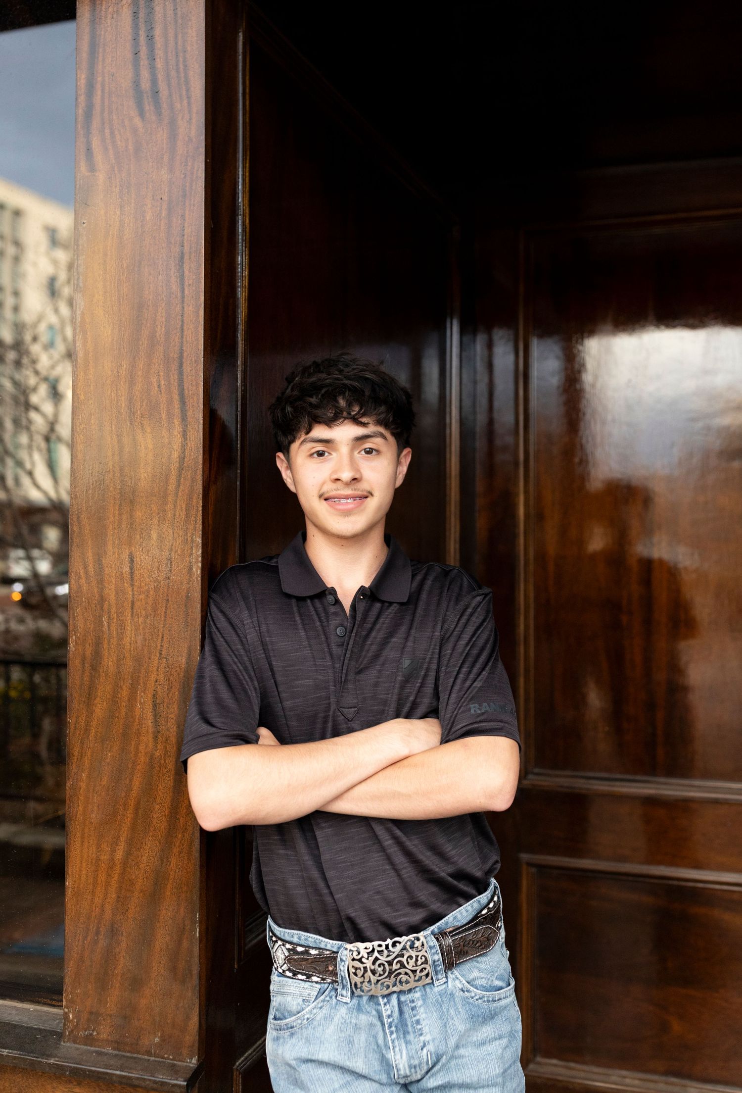 Young man with crossed arms smiling while leaning against wooden doorframe outdoors.