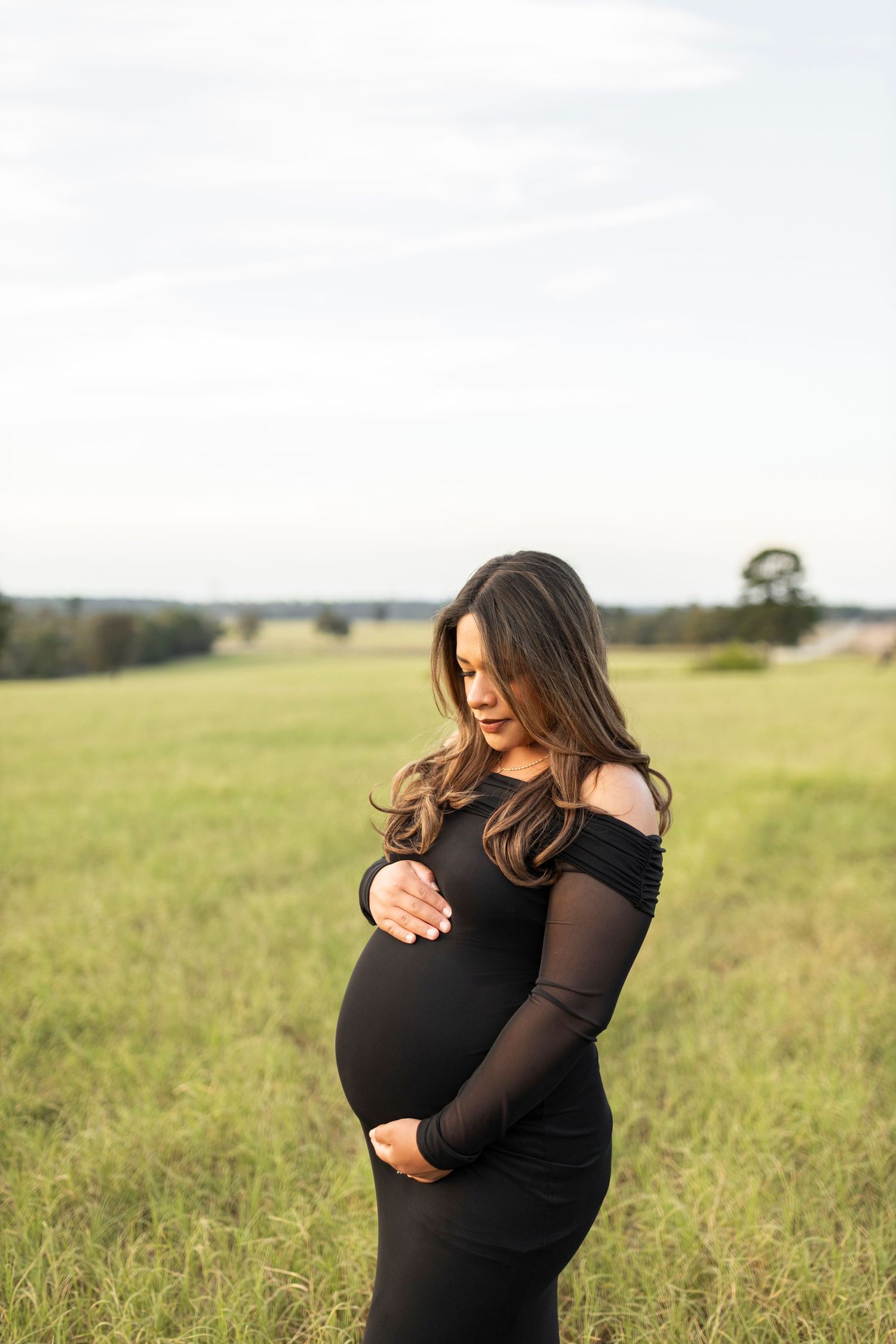 Pregnant woman in black off-shoulder gown cradling her belly in an open green field.