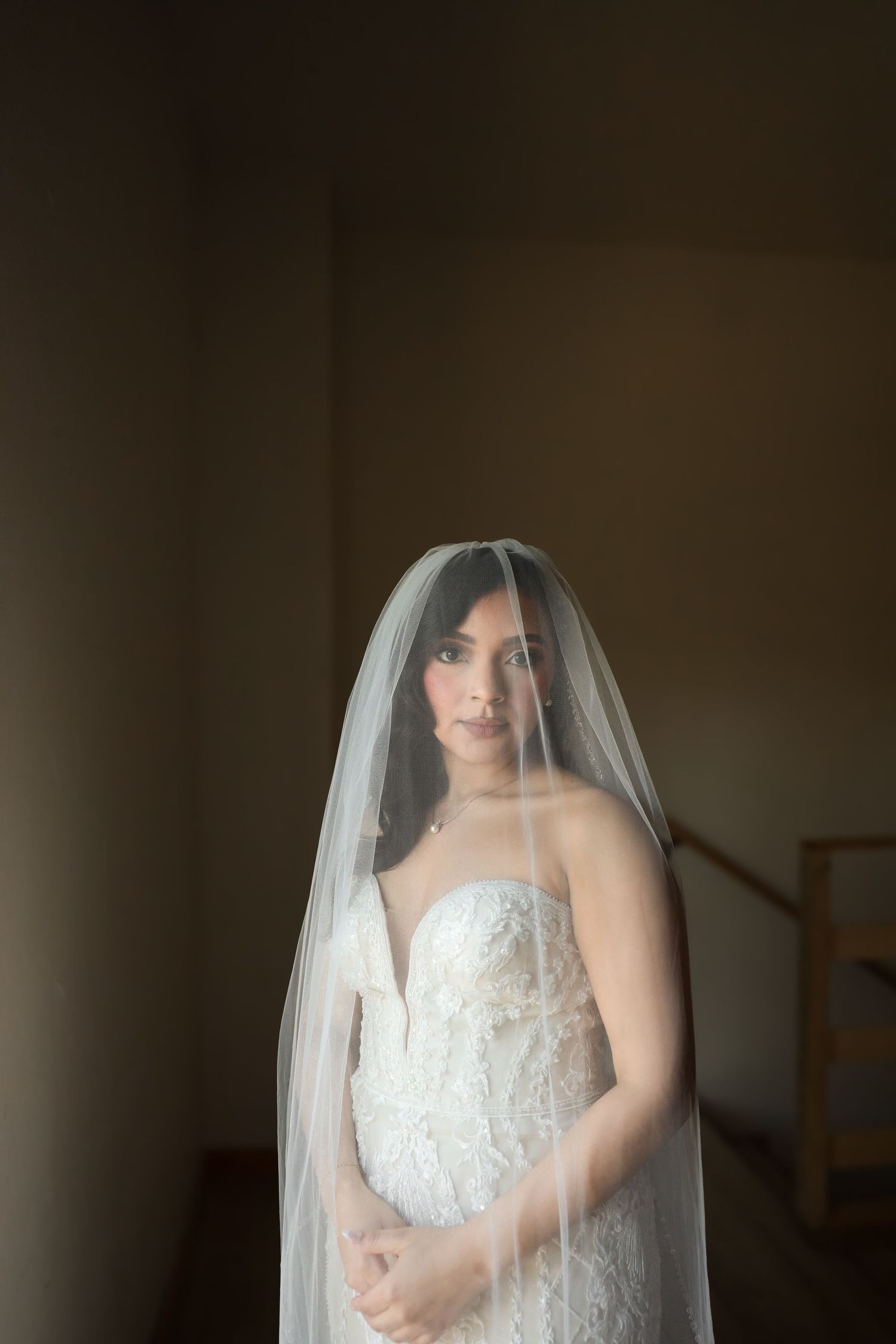 Elegant bride in lace strapless gown with cathedral veil gazing forward in softly lit interior.
