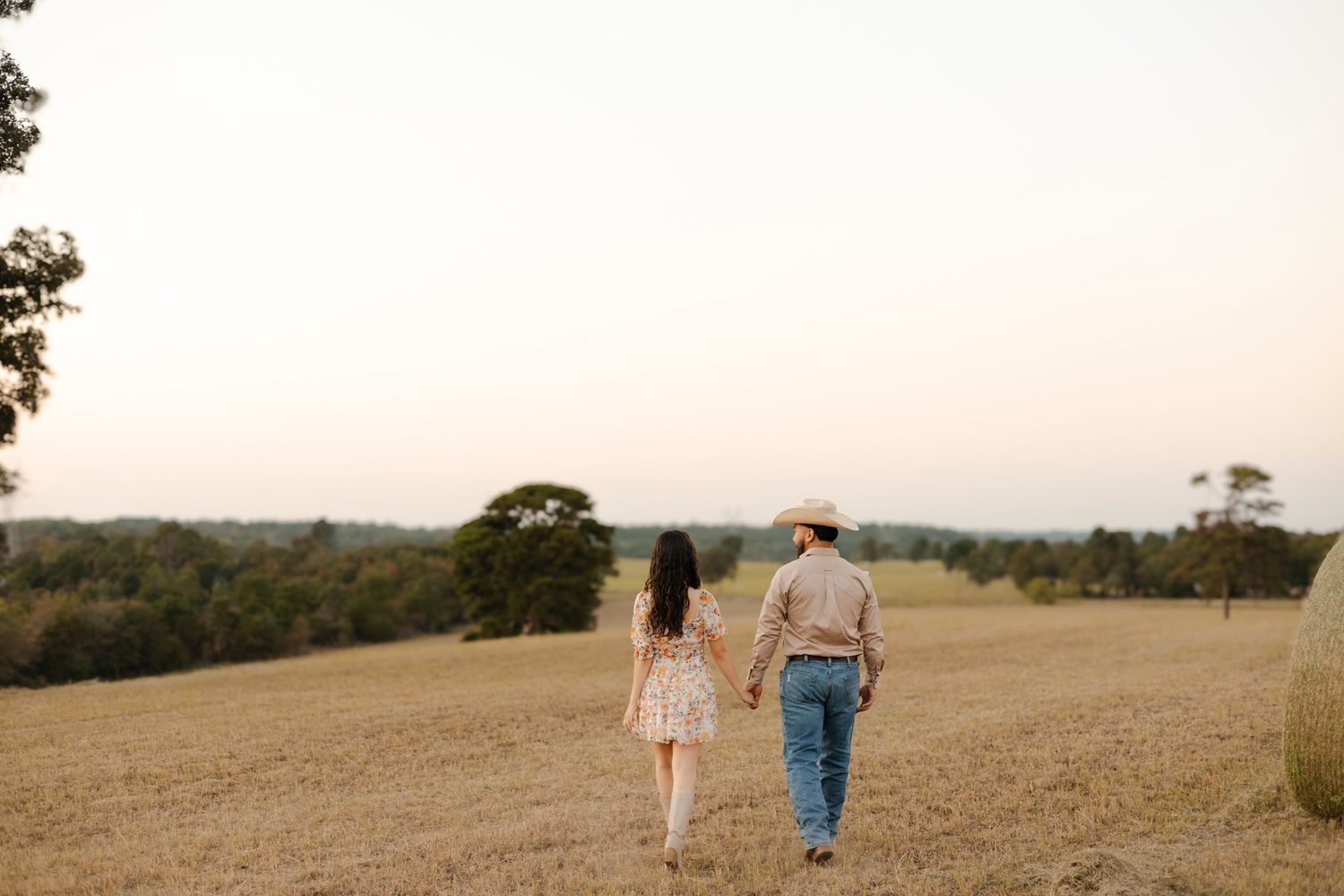 Couple holding hands walking through a golden harvest field at sunset in country attire.