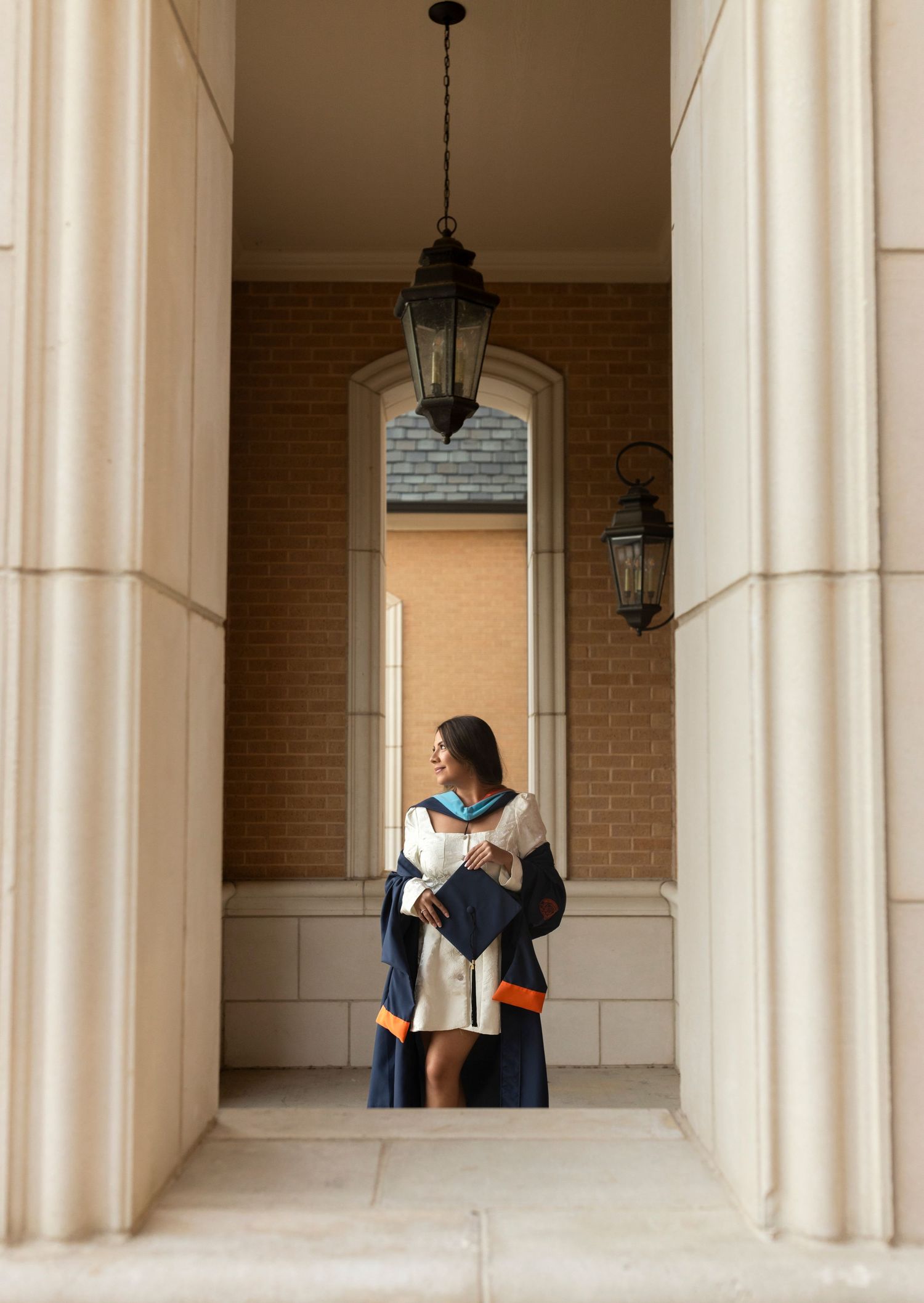 Graduate in navy and orange regalia stands between tall stone columns with hanging lantern above.