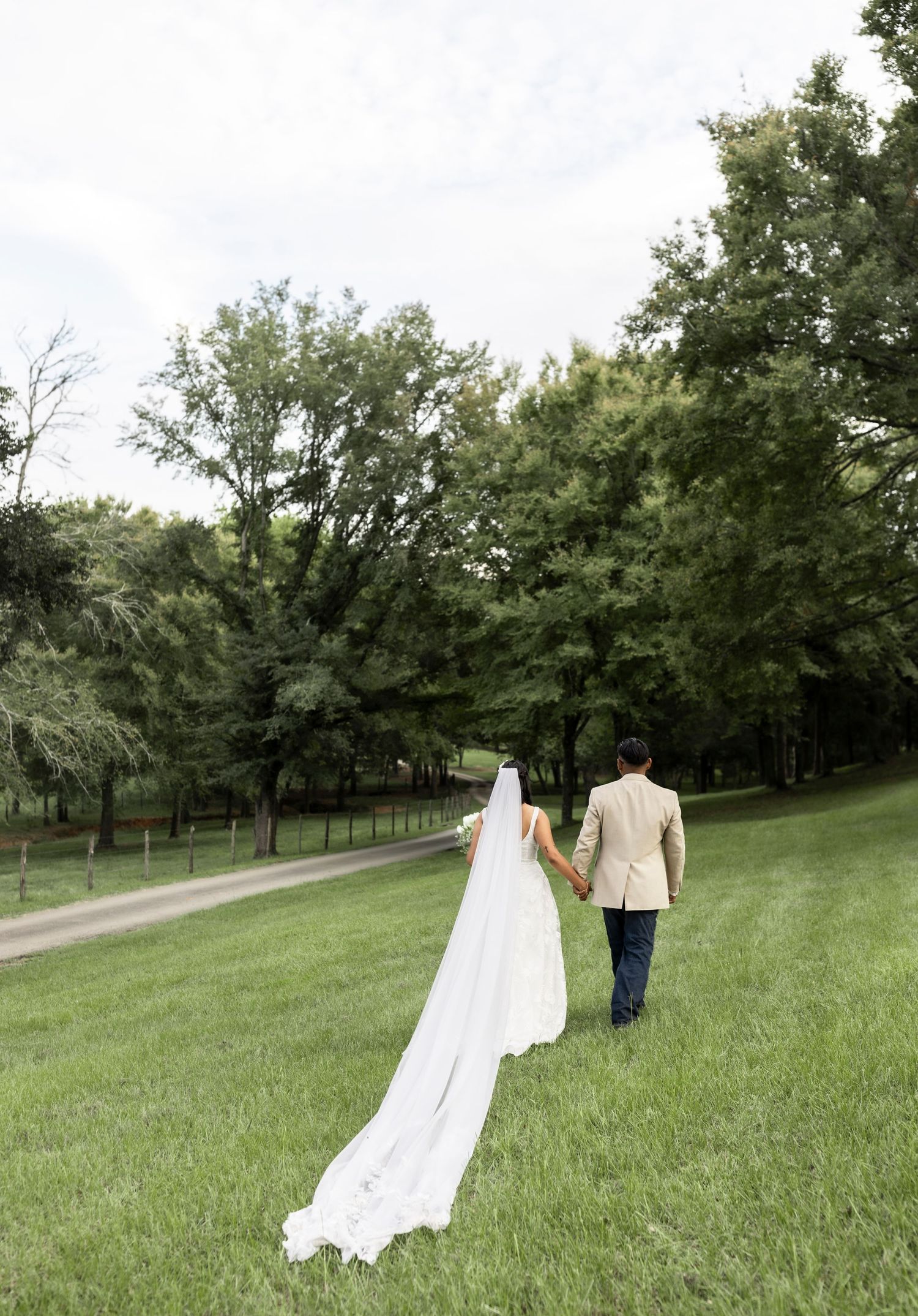Bride and groom walk hand-in-hand across a lush green lawn, her cathedral veil trailing behind.