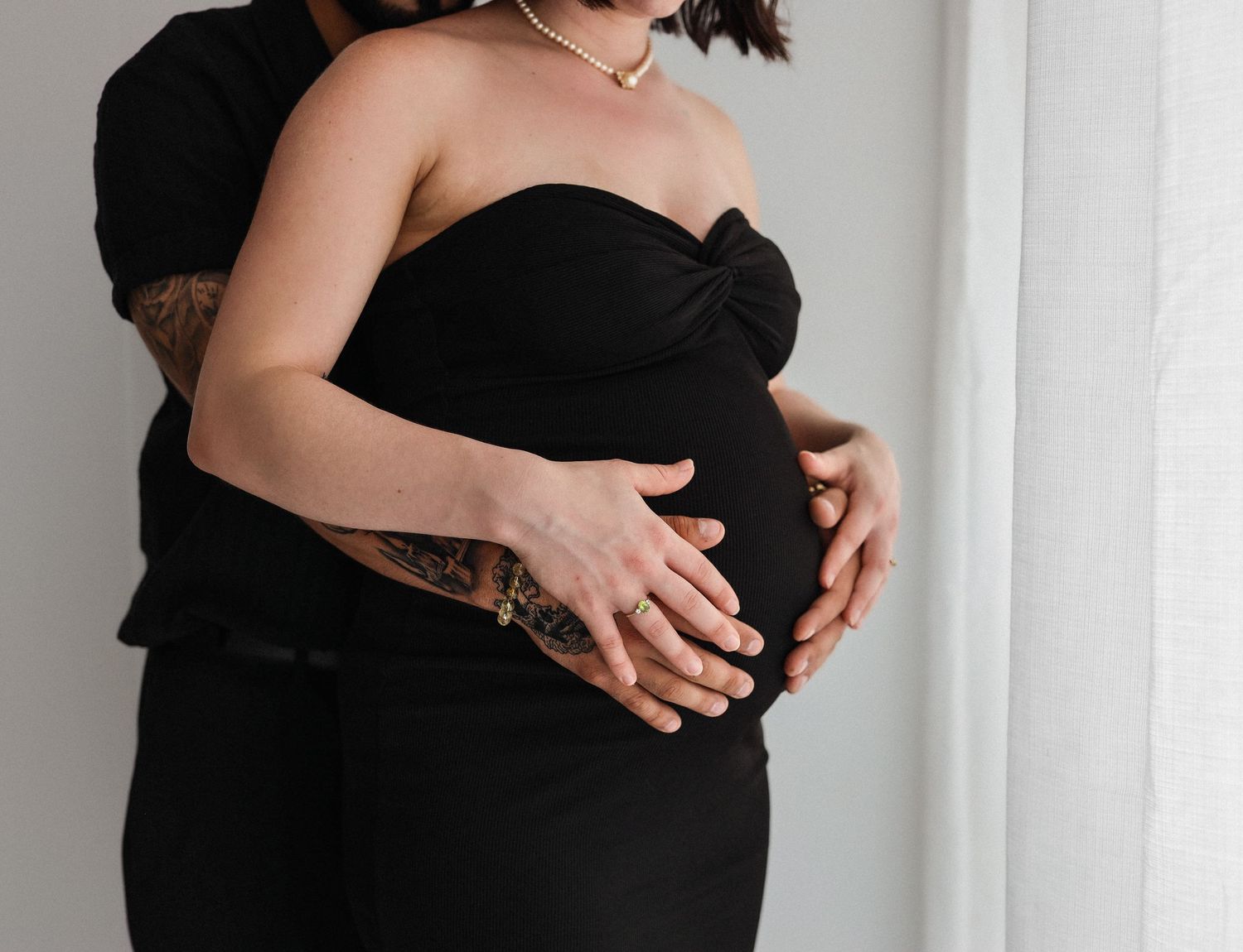 Close-up of pregnant couple in black outfits, tattooed partner embracing baby bump together.