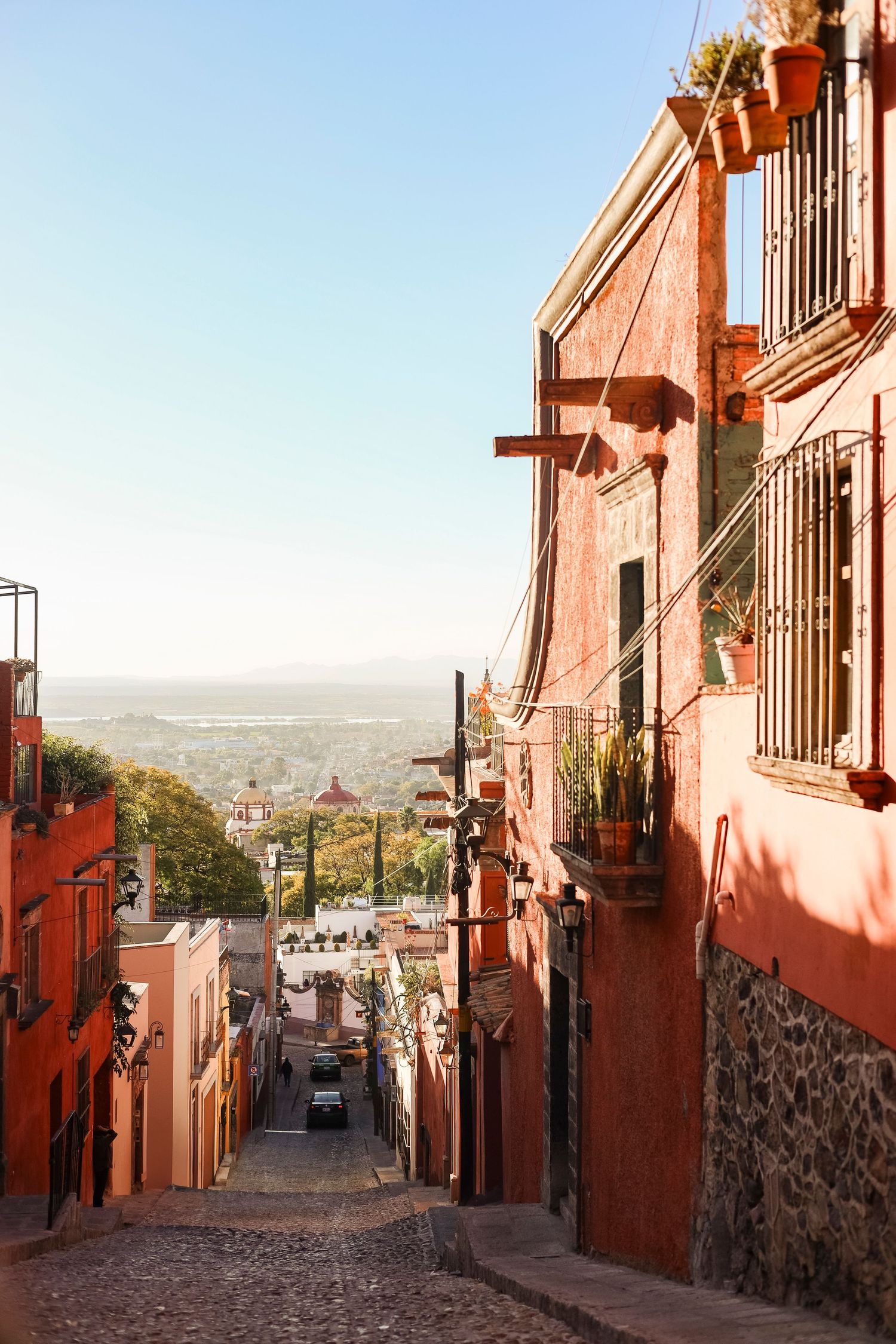 Colorful cobblestone street in San Miguel de Allende lined with terracotta buildings and city views below.