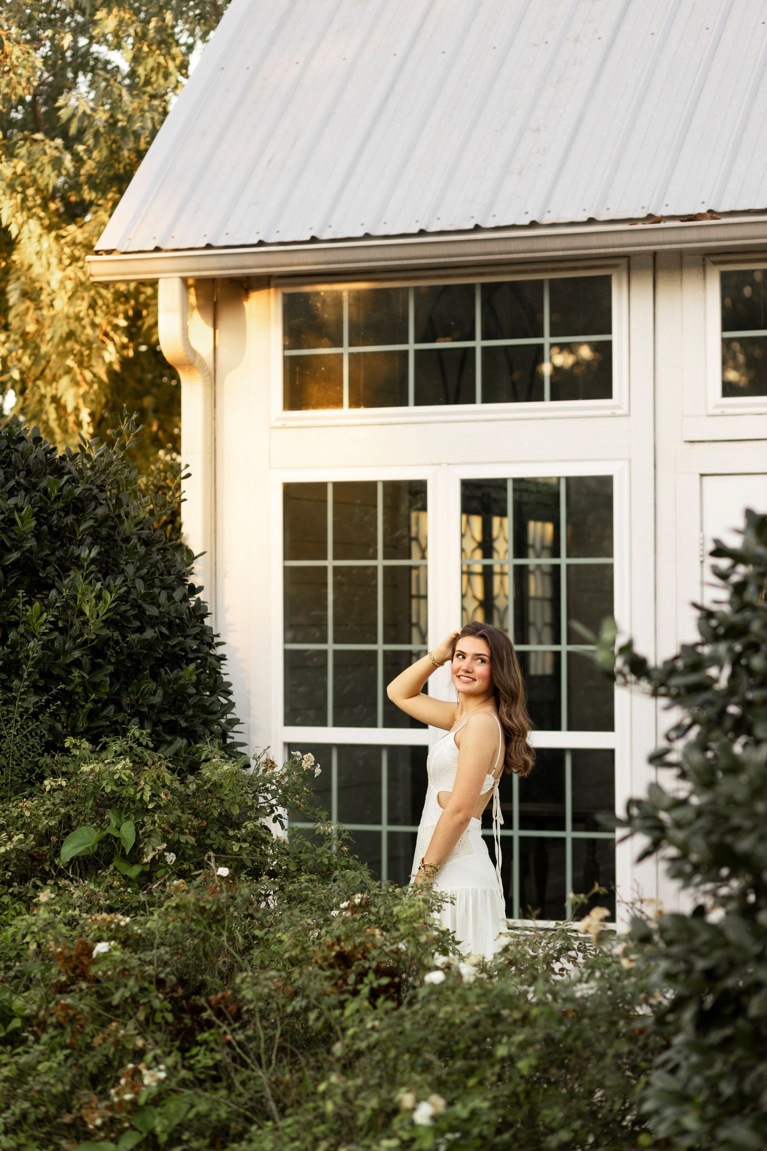 Woman in white dress stands among lush garden greenery before white building with large windows at golden hour.