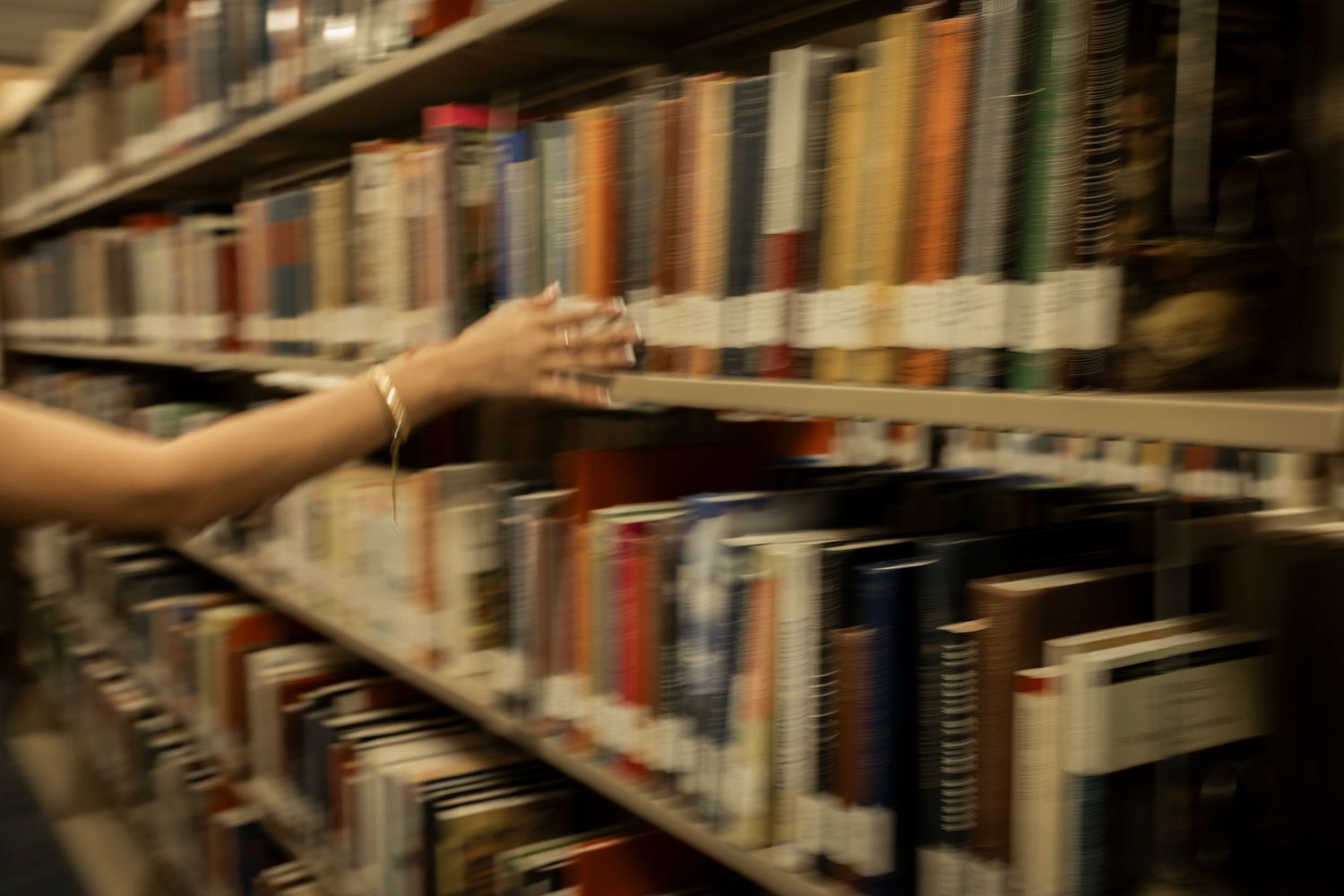 Close-up of hand reaching for books on a dimly lit library bookshelf.
