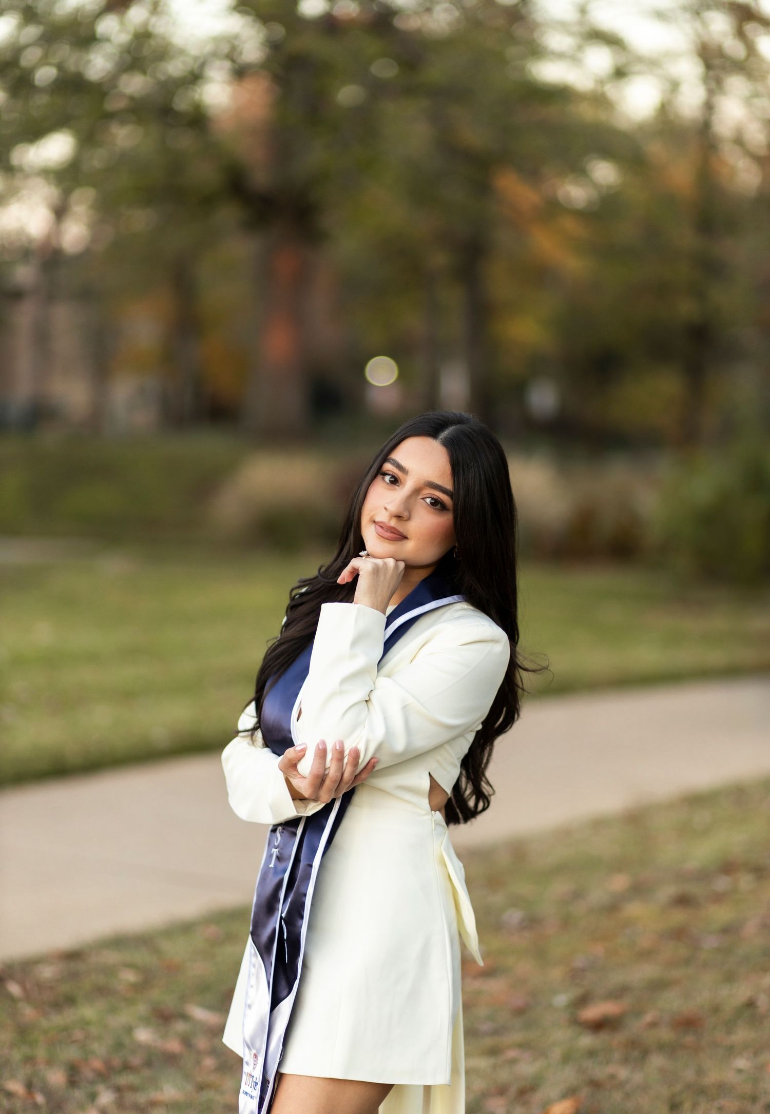 Female graduate in white dress with navy sash poses gracefully outdoors in autumn setting.