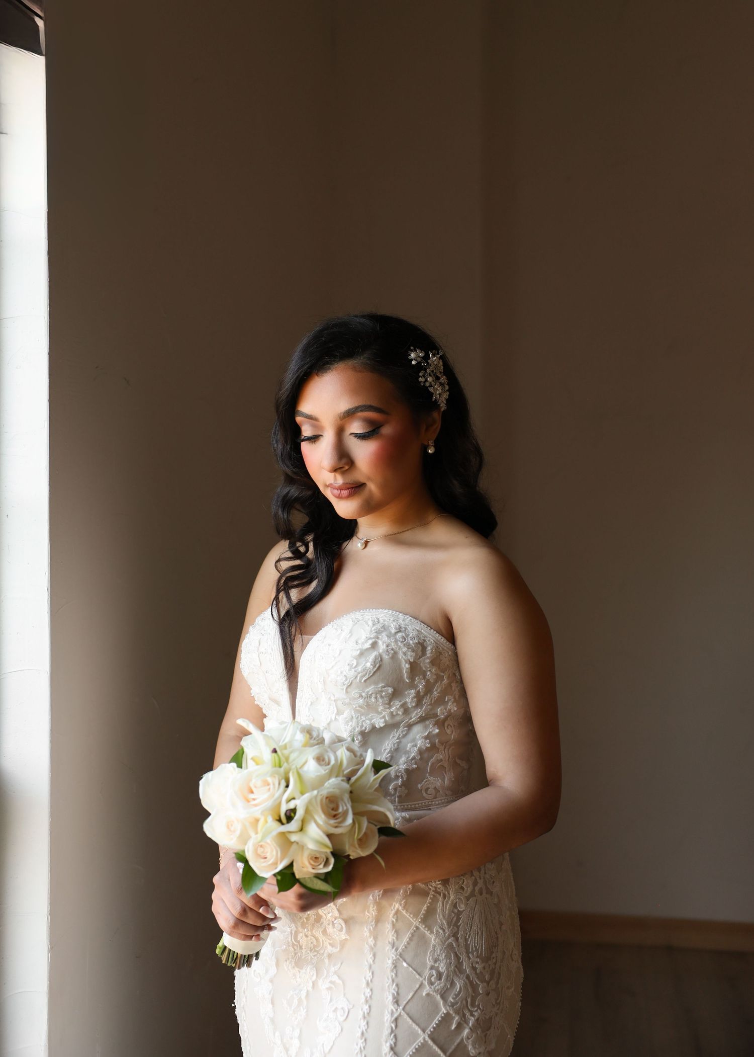 Bride in strapless lace gown holding white rose bouquet with floral hair piece near window light.