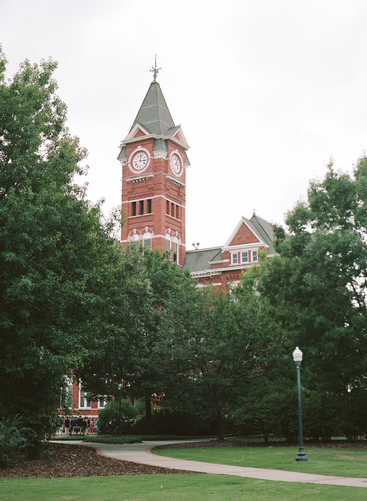 Historic red brick church building with clock tower and spire surrounded by green trees.