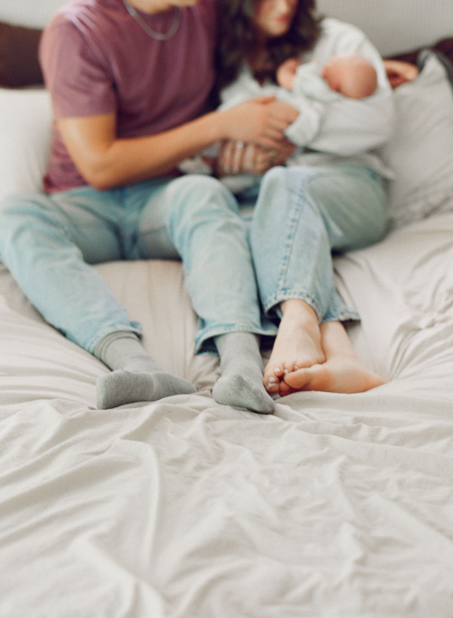 A couple sits on a white bed holding their newborn baby, wearing jeans, captured in a cozy intimate family photo.