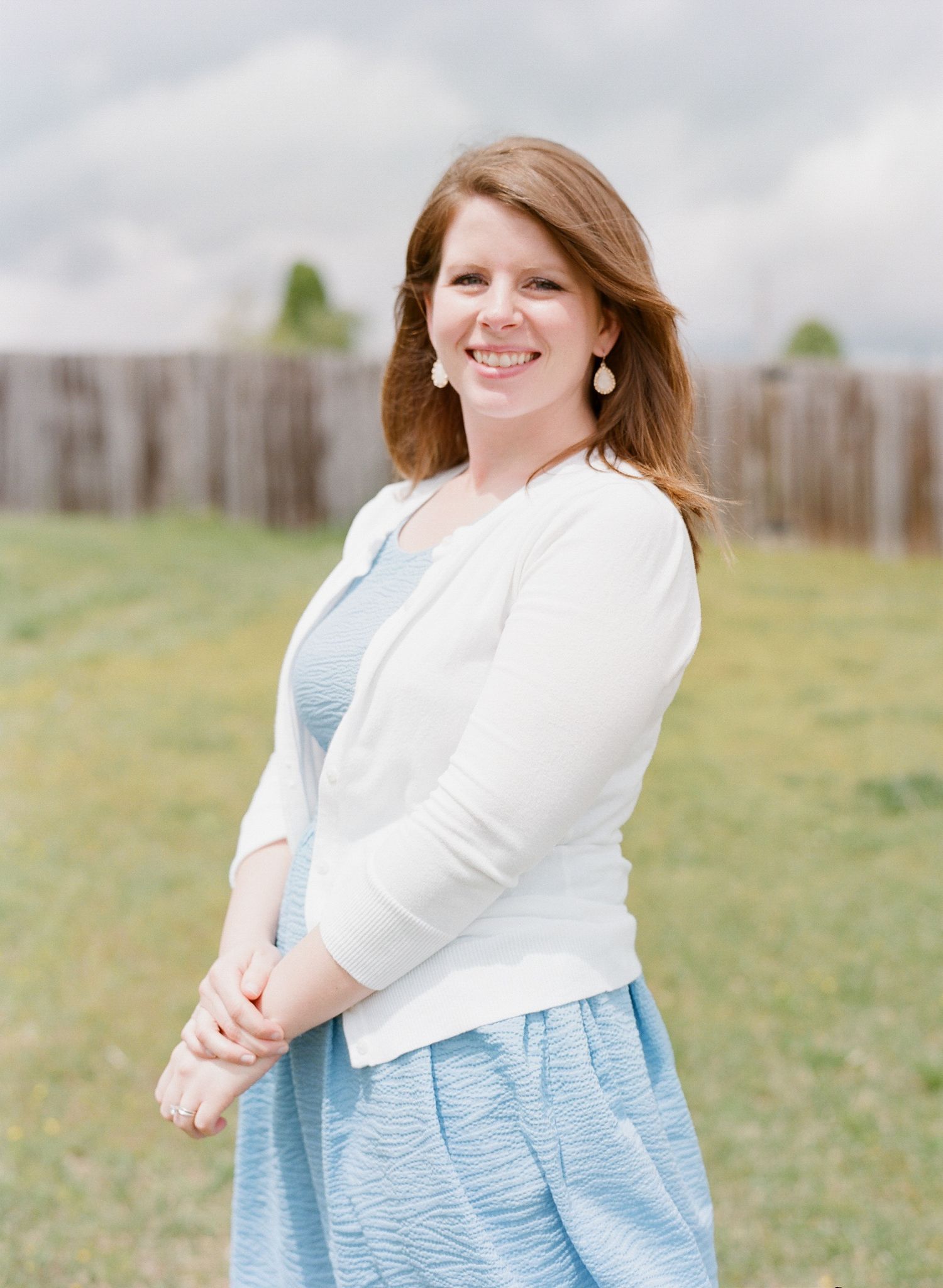 Portrait in a casual white top and blue skirt against an outdoor backdrop with a wooden fence.