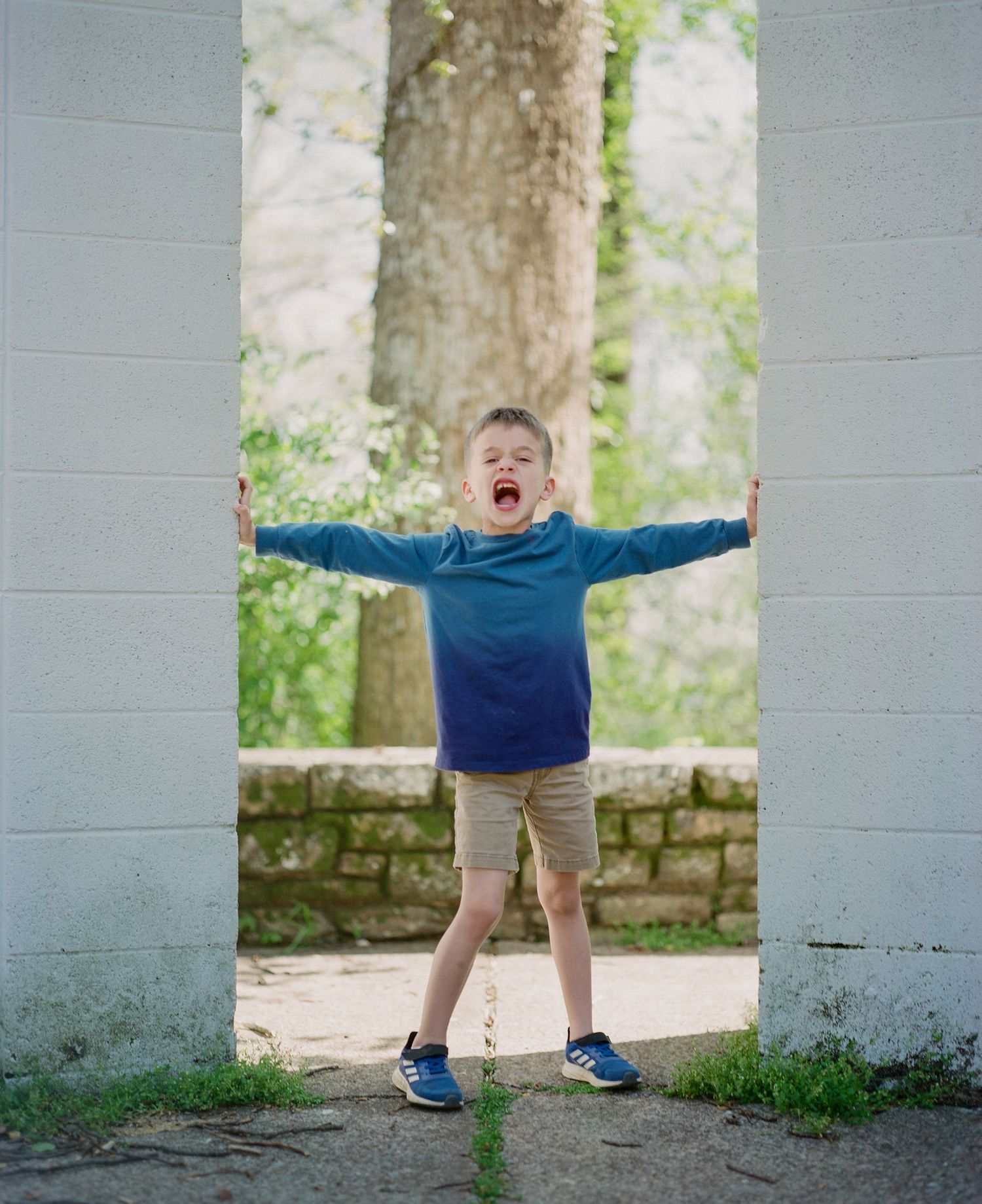Child in navy shirt and khaki shorts expressing joy with arms raised outdoors.