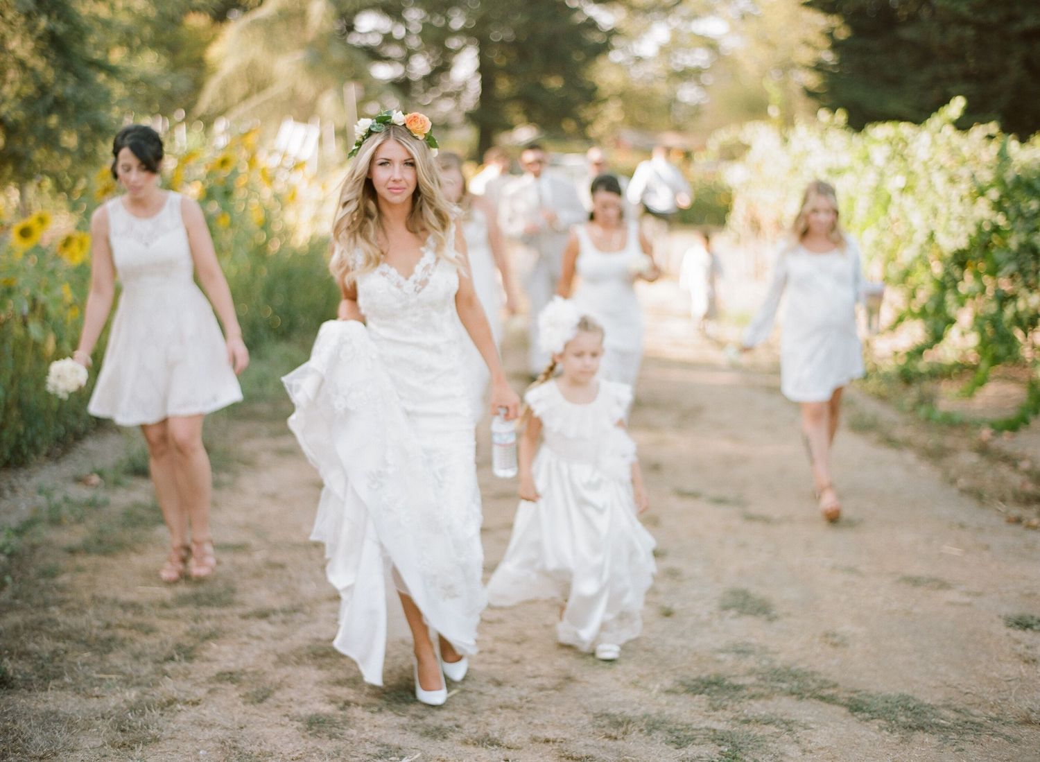 Wedding party in white dresses walks along a sunlit outdoor path.