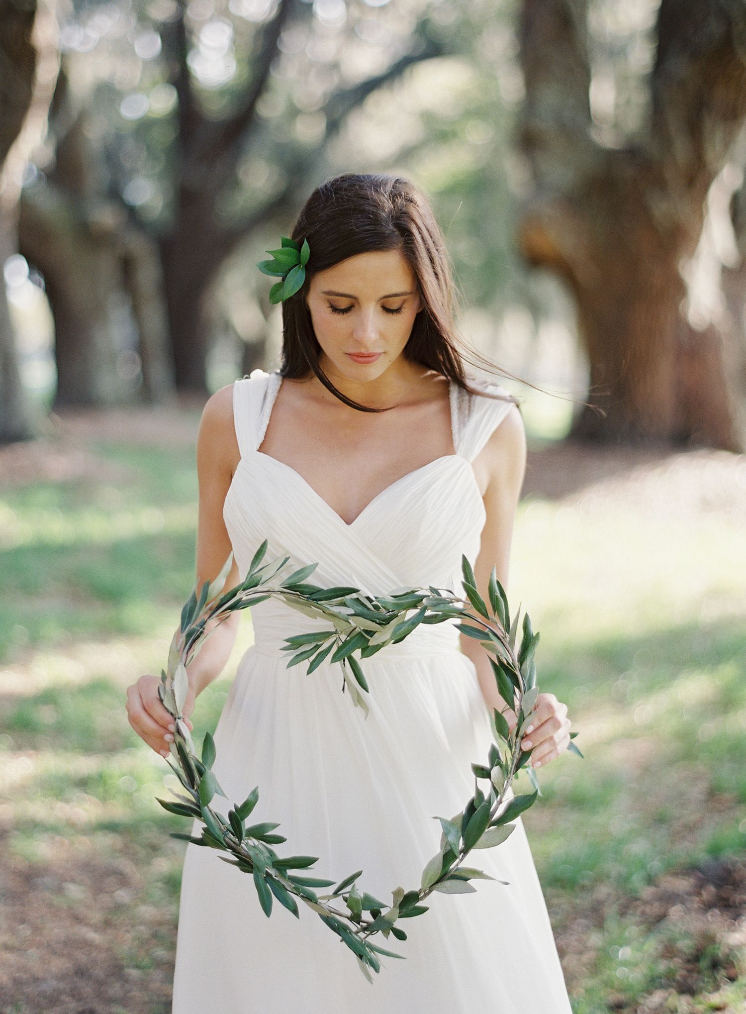 Bride in white dress holding olive branch wreath in a sun-dappled grove.