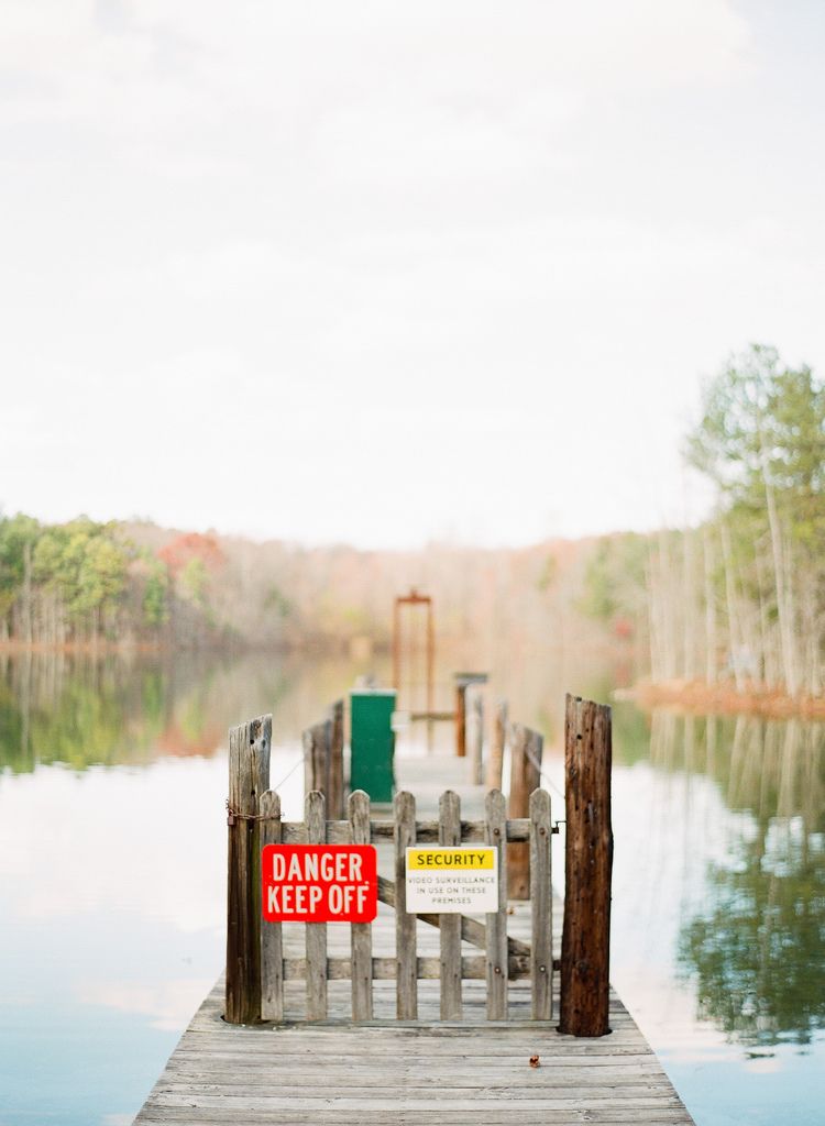 Wooden dock with danger signs extending into a serene lake surrounded by autumn trees.