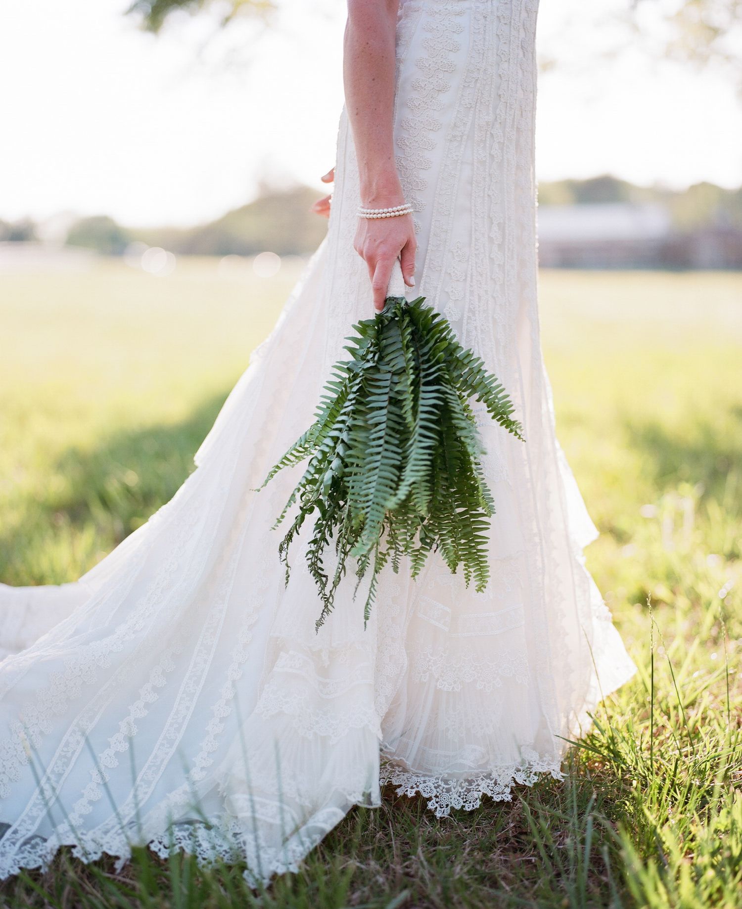 Close-up of a white lace wedding dress hem with fern bouquet against sunlit grass.