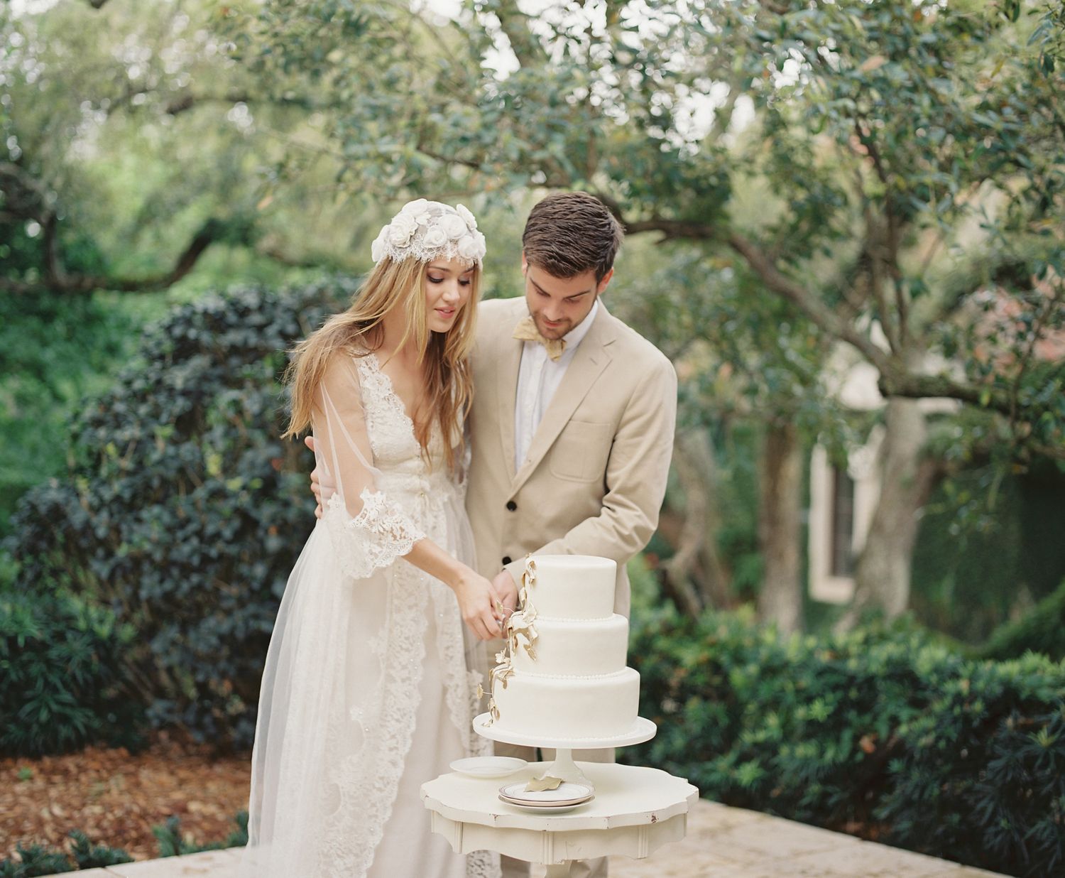 Couple in wedding attire cutting their white cake together in an outdoor garden setting.