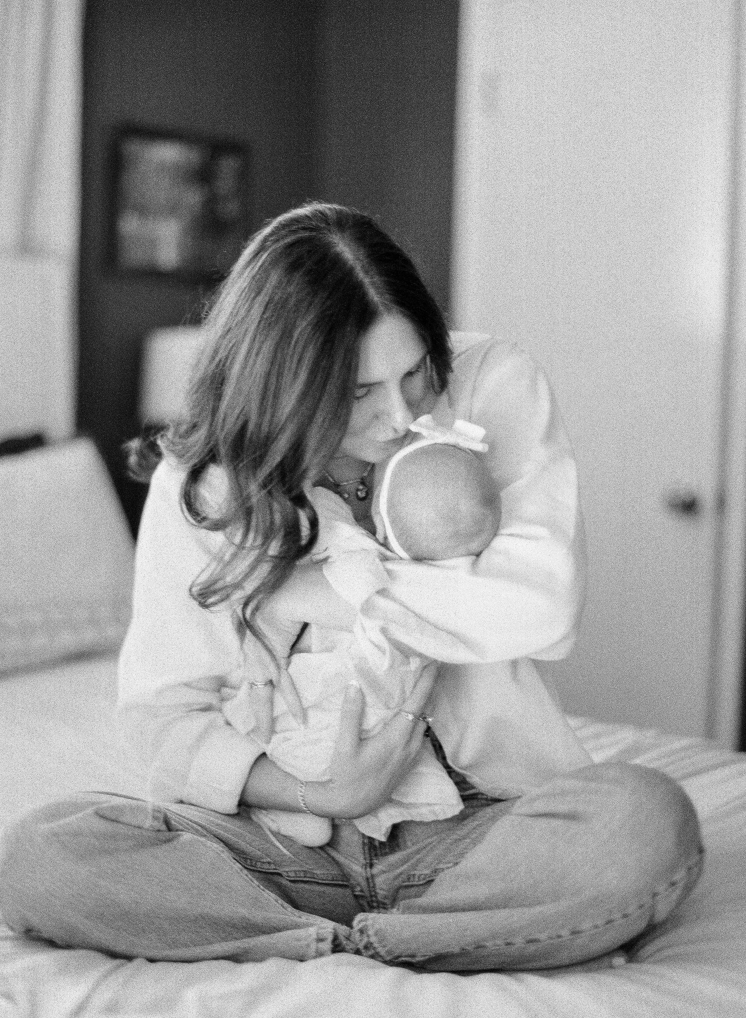 Black and white photo of a mother sitting cross-legged on a bed, tenderly kissing her newborn baby.