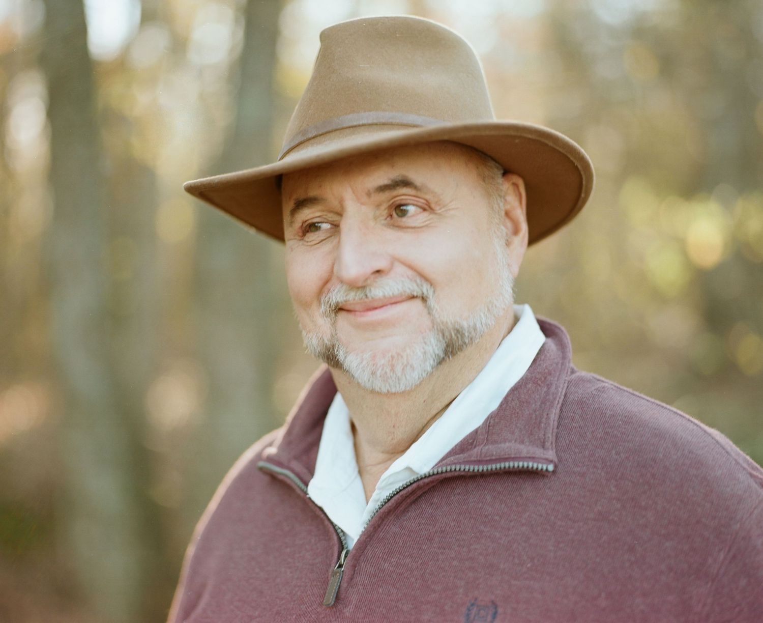 Side profile of a man in brown hat and burgundy jacket against blurred forest background.