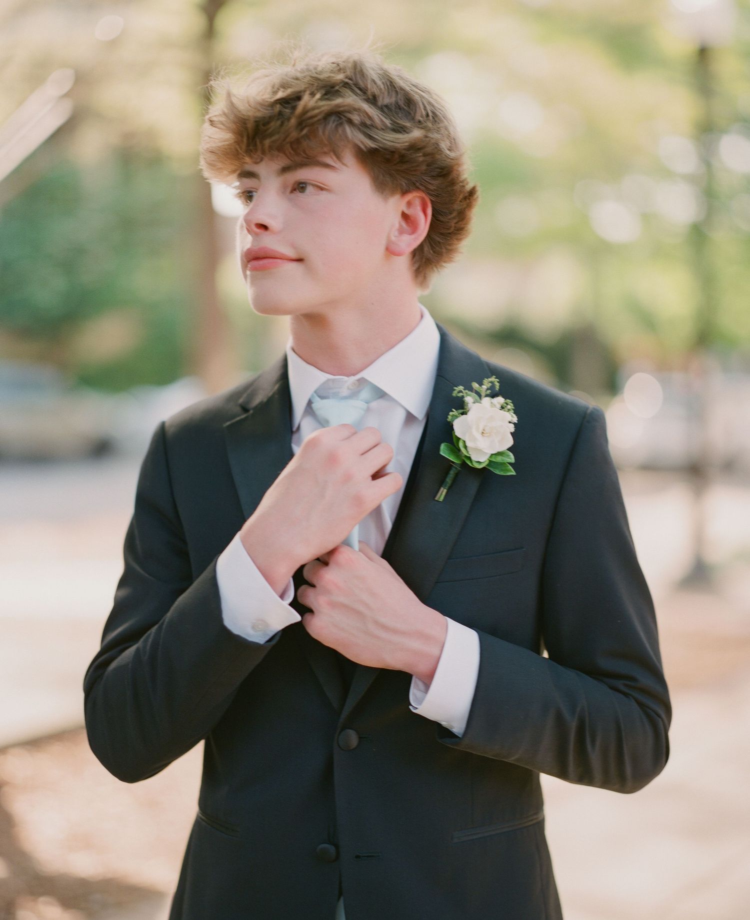 Young man in dark suit adjusting tie with white boutonniere, elegant formal attire for special occasion.